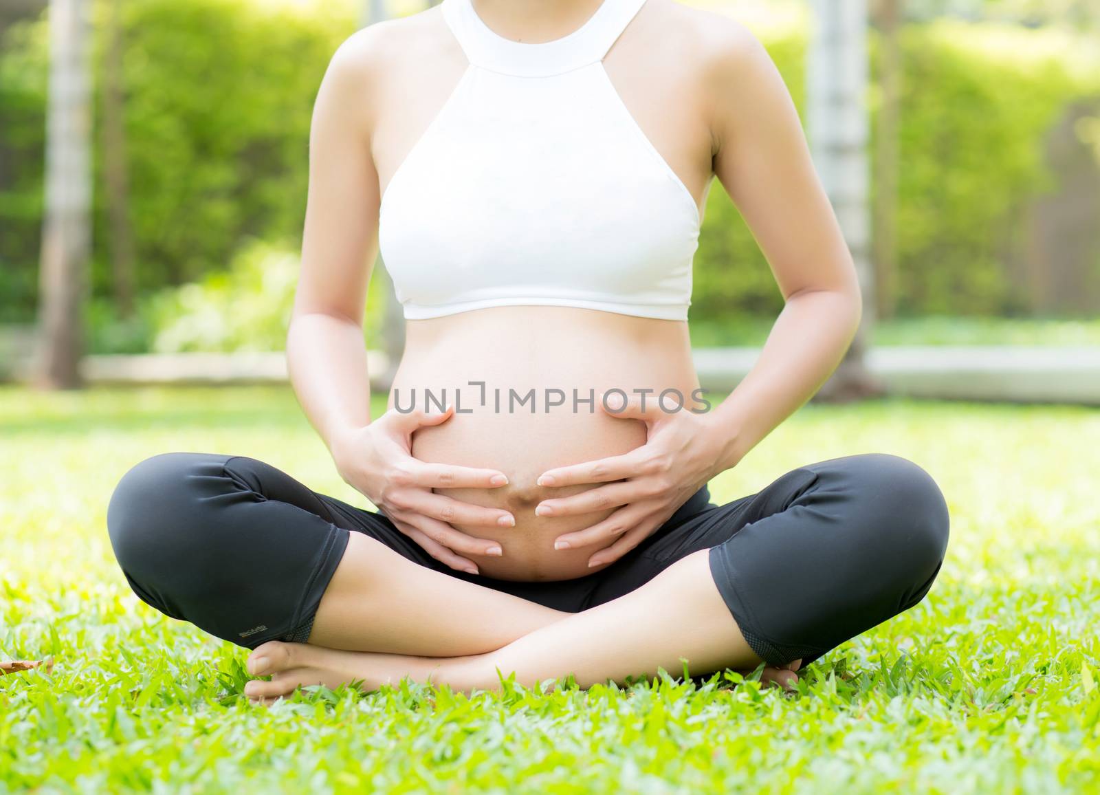 Beautiful pregnant asian young woman relax in the park, girl with belly sitting with crossed leg on grass and yoga exercise for wellness, copy space.