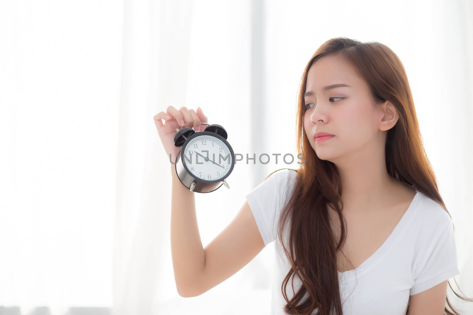 Beautiful young asian woman wake up in morning annoyed alarm clock holding hand, girl standing hurry wake late with appointment with curtain background on bedroom, lifestyle concept.