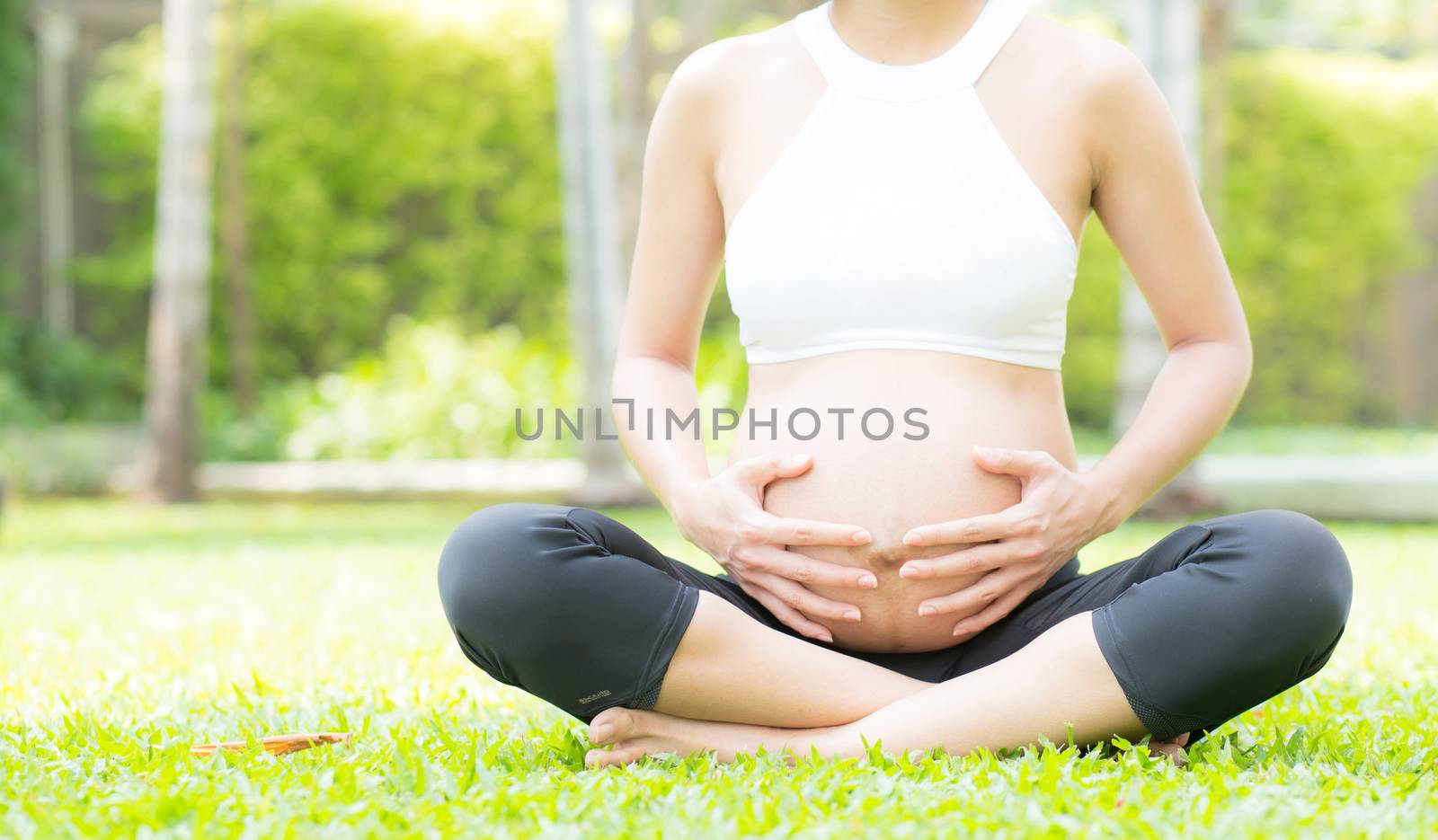 Beautiful pregnant asian young woman relax in the park, girl with belly sitting with crossed leg on grass and yoga exercise for wellness, copy space.