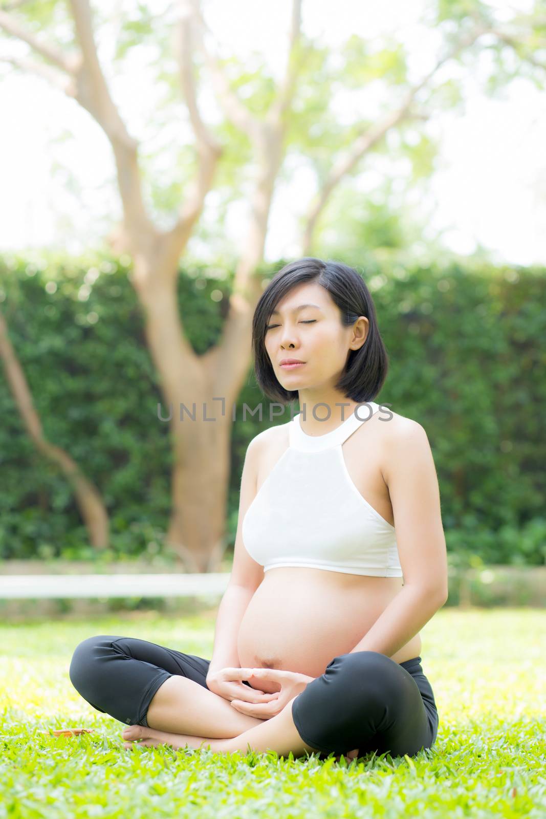 Beautiful pregnant asian young woman relax in the park, girl with belly sitting with crossed leg on grass and yoga exercise for wellness, copy space.