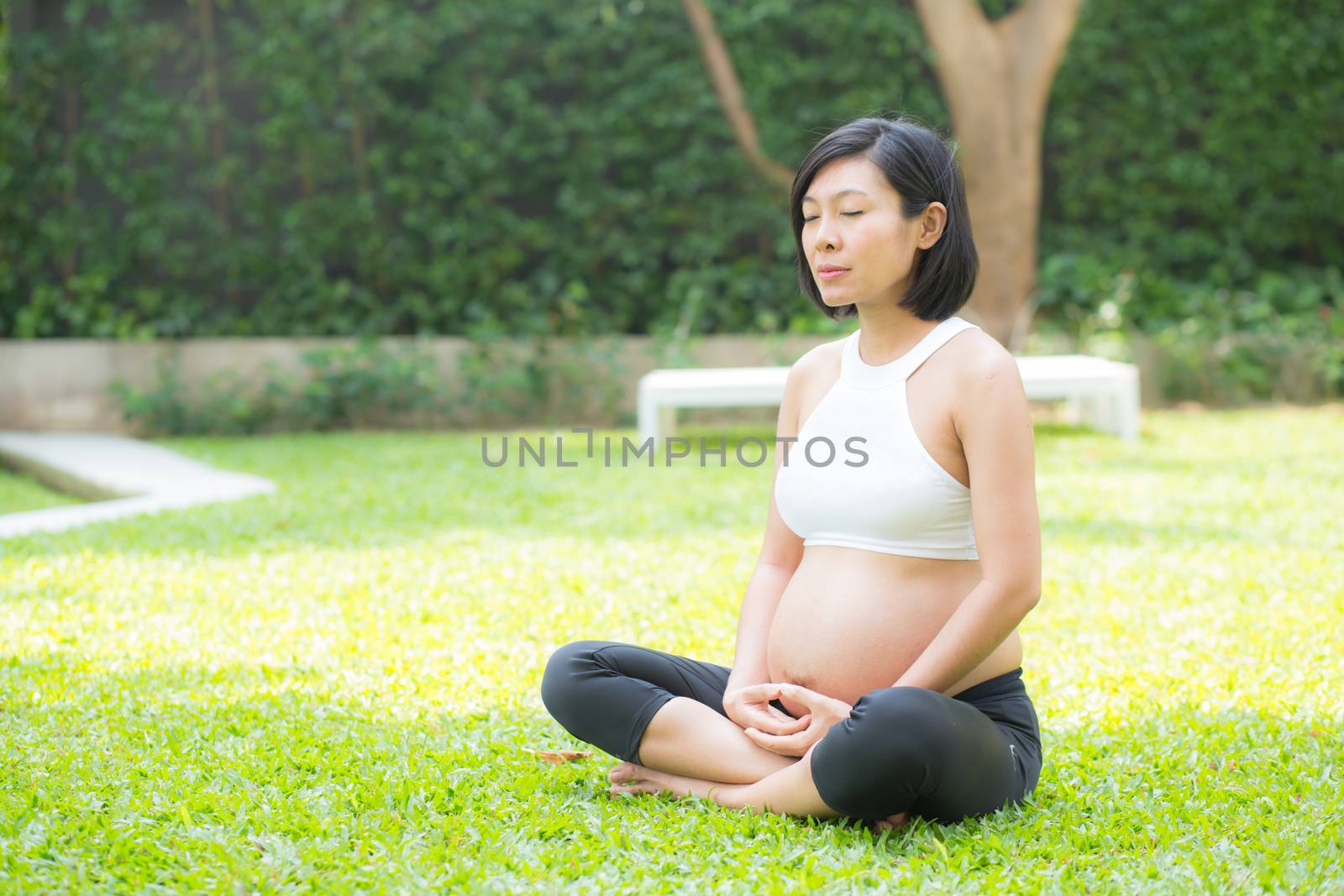 Beautiful pregnant asian young woman relax in the park, girl with belly sitting with crossed leg on grass and yoga exercise for wellness, copy space.