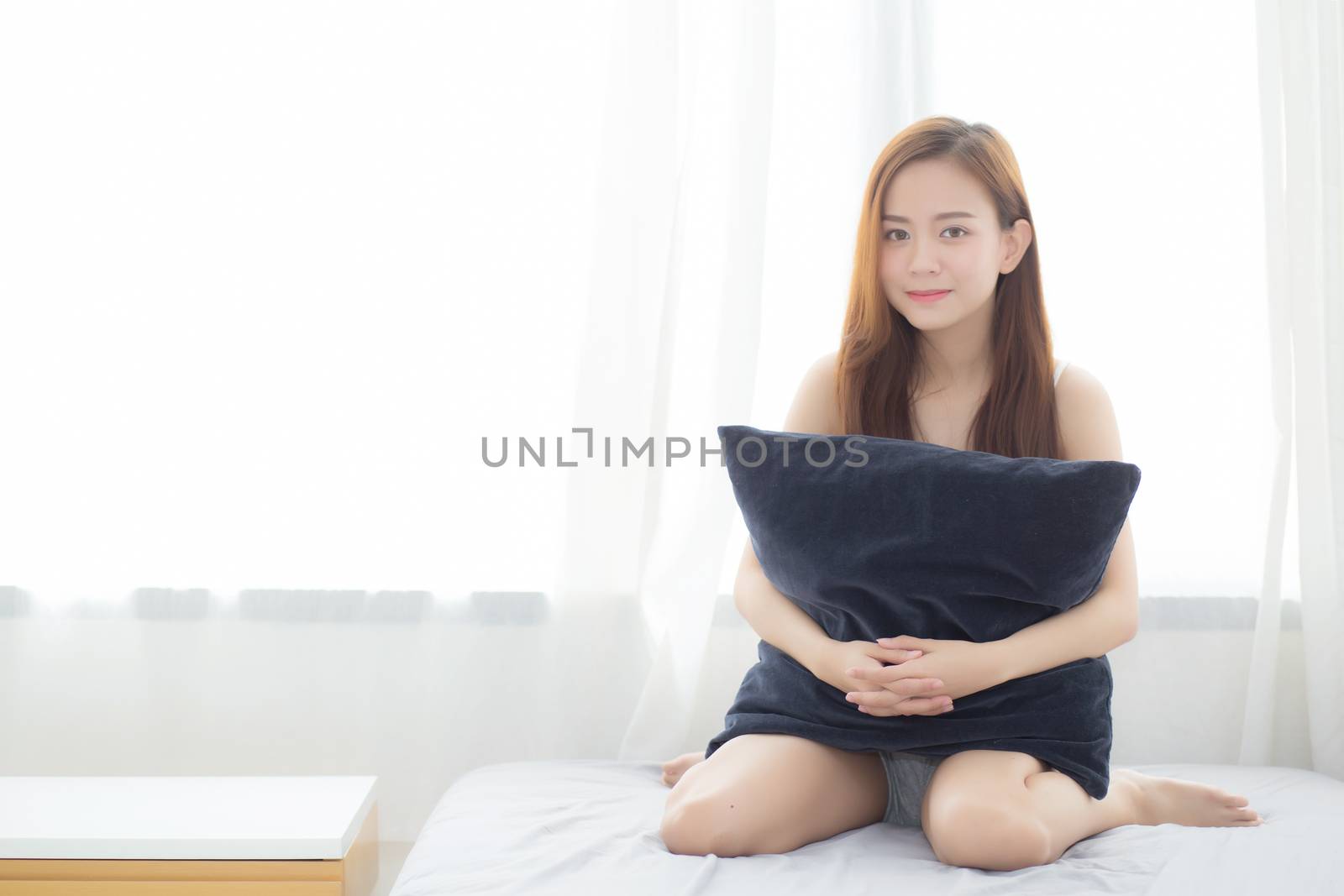 Portrait beautiful young asian woman sitting and smile hug pillow with the window at bedroom while wake up with sunrise at morning, lifestyle and relax concept.