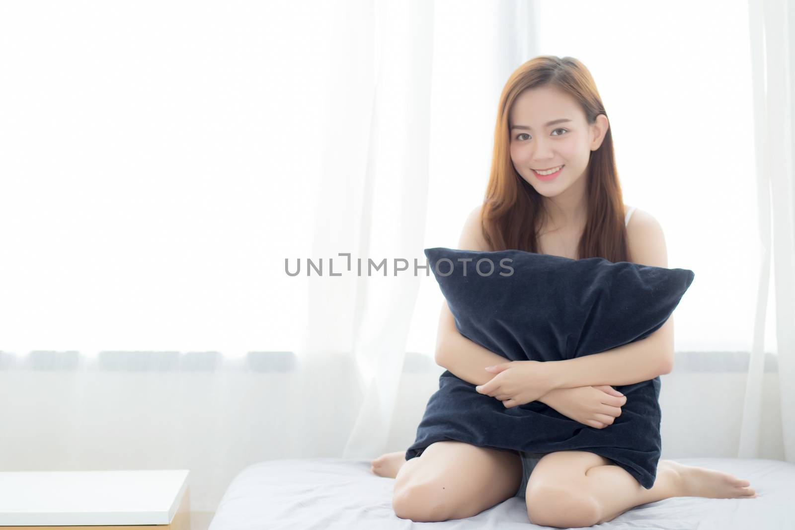 Portrait beautiful young asian woman sitting and smile hug pillow with the window at bedroom while wake up with sunrise at morning, lifestyle and relax concept.