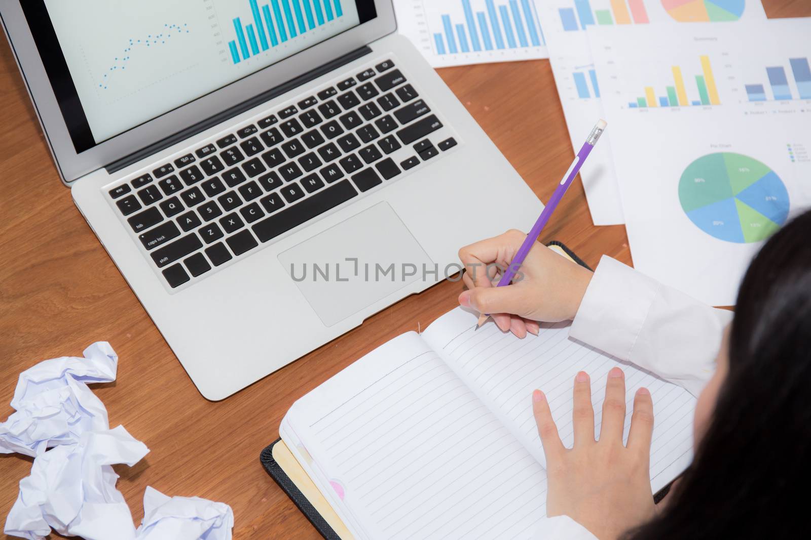 Businesswoman writing on notebook at desk, asian woman with not idea with graph analysis laptop and crumpled paper at office, freelance business concept.