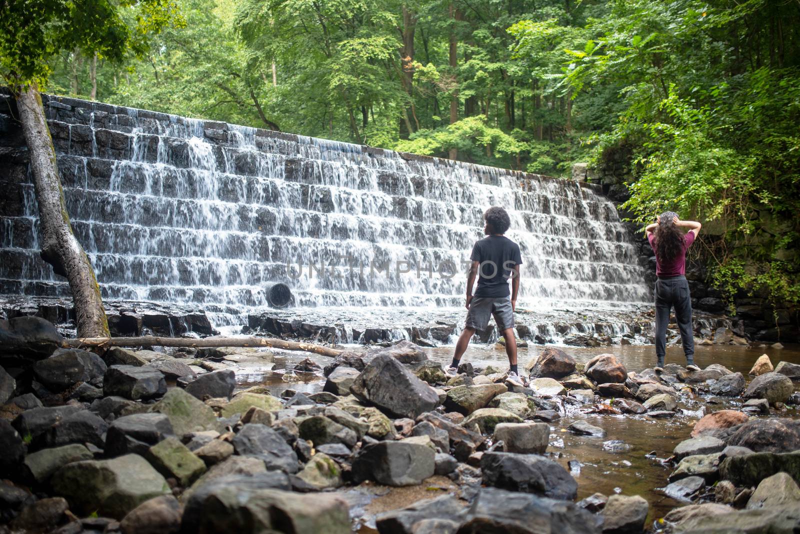 Water cascades over the tiered waterfall with lush woodland foliage behind. Two young men, African-American and Caucasion, stand on stones at base of falls.