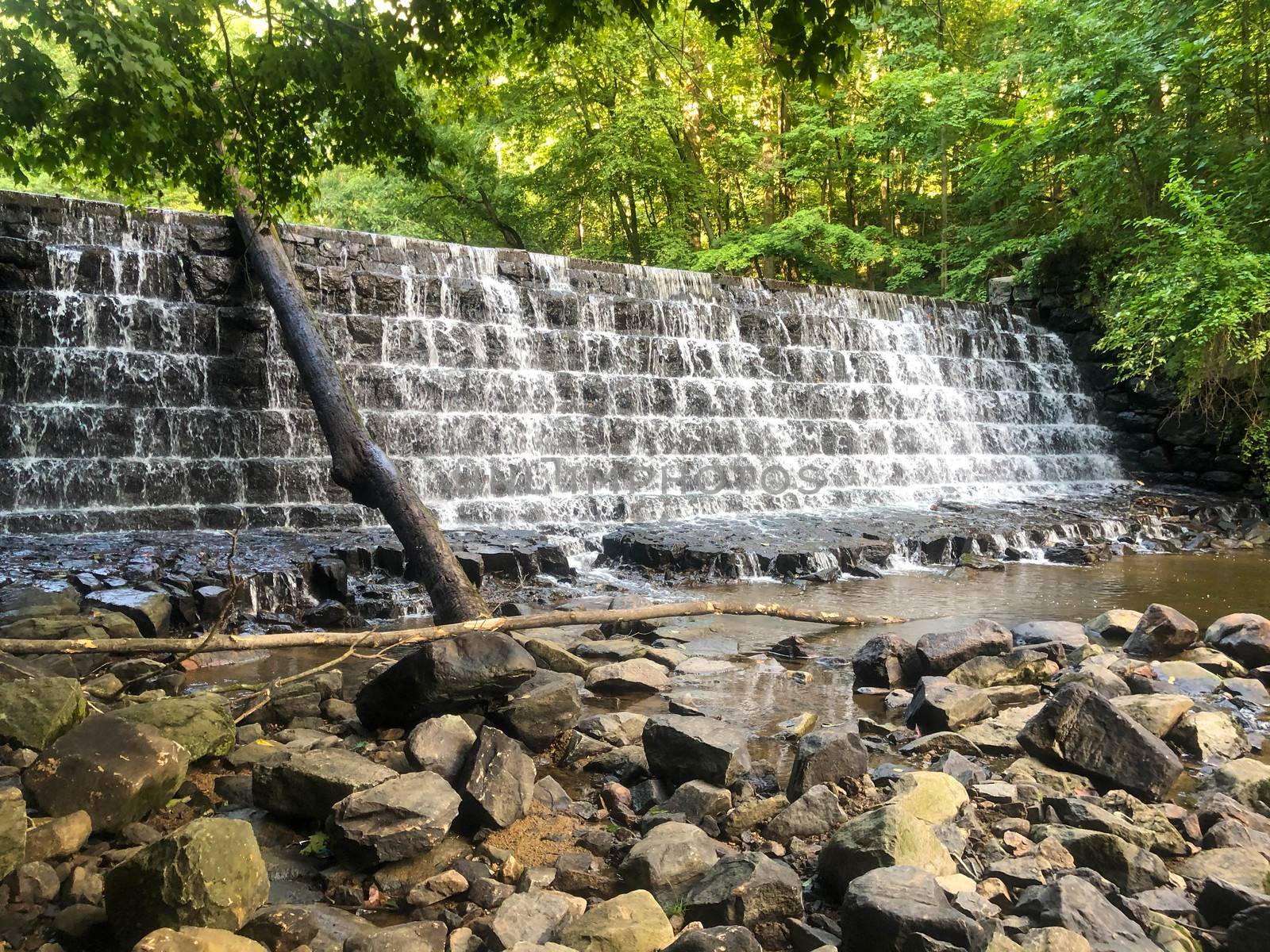 Beautiful scenic image of a tiered waterfall of rough textured stones. Green woodland in background and river stones in foreground. Excellent nature background with copy space.