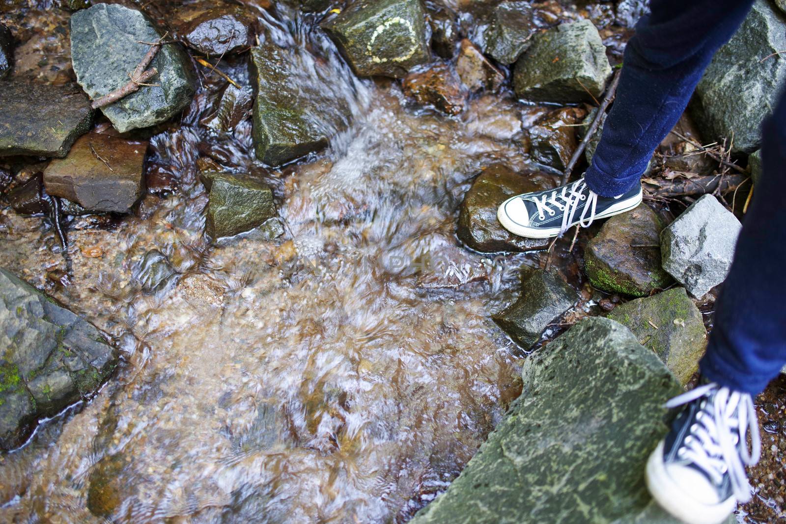 A young man in blue pants and sneakers climbs across a flowing stream on colorful stepping stones. Natural light with copy space.