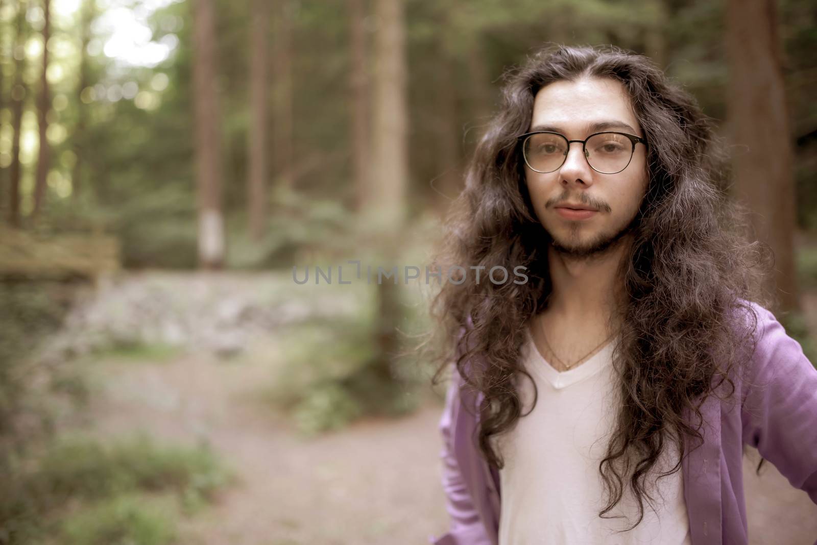 A handsome young man with long hair stands off center in the woods, looking straight at the camera. Defocused background copy space.