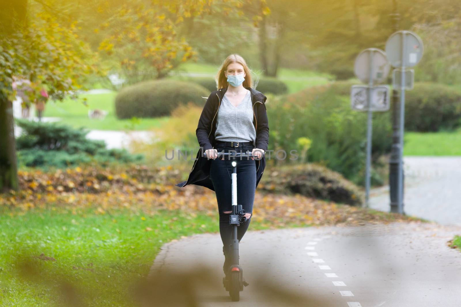 Casual caucasian teenager wearing protective face mask riding urban electric scooter in city park during covid pandemic. Urban mobility concept.