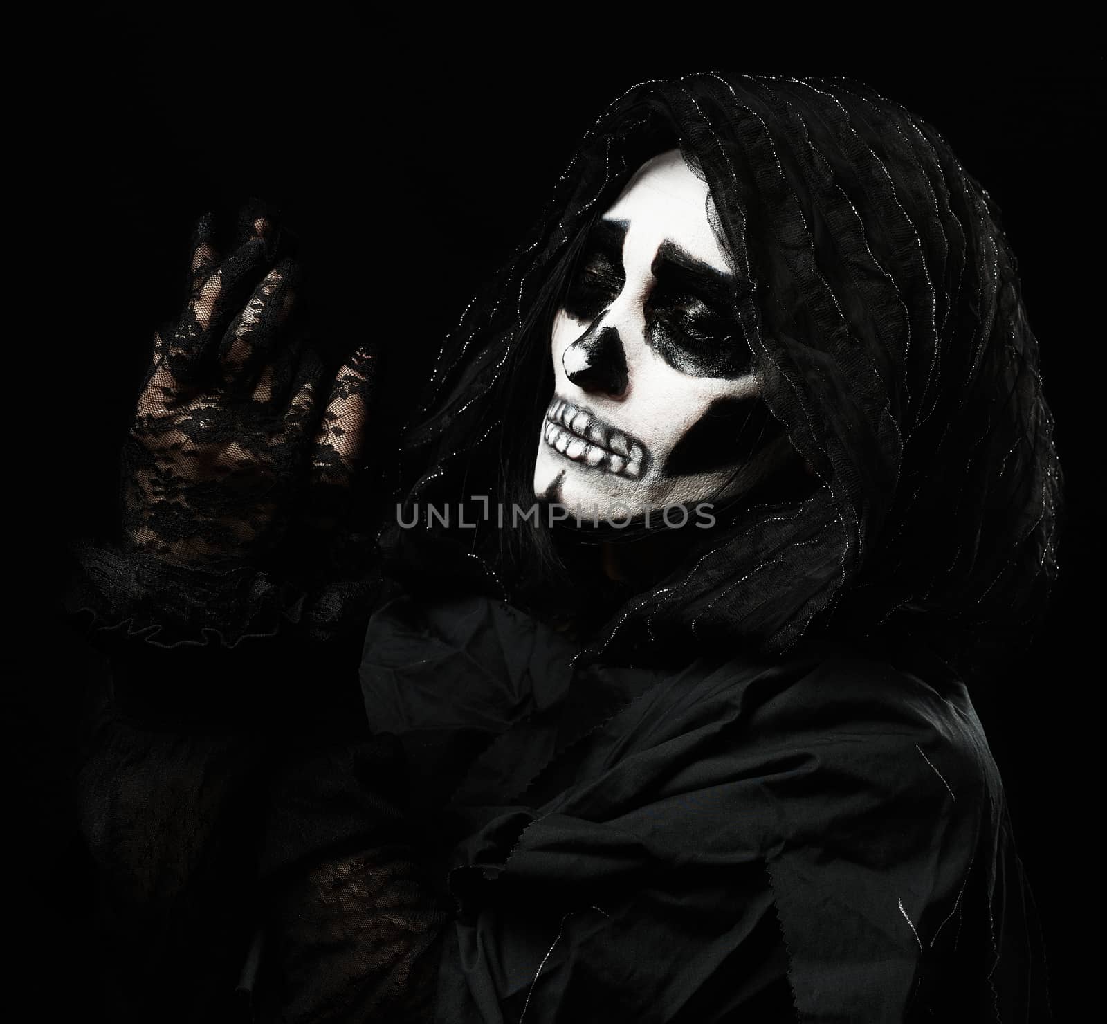 woman with a make-up skeleton stands in black clothes and a transparent hood, hands raised up, black background