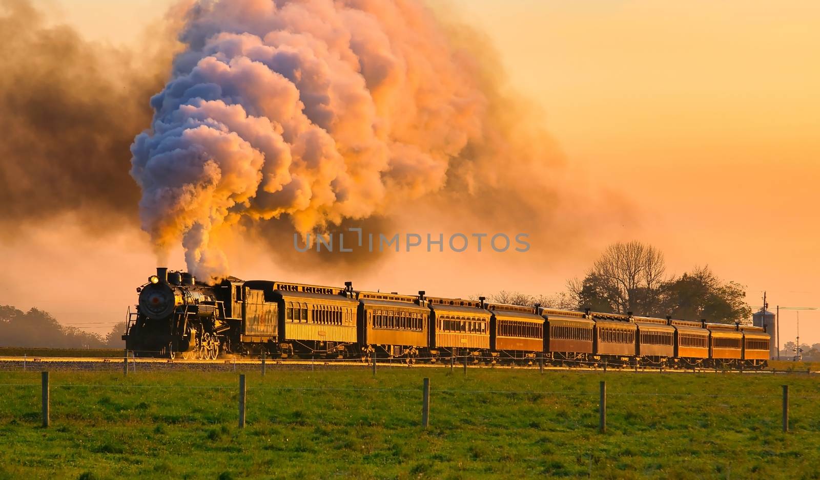 View of Golden Steam Passenger Train at Sunrise Traveling Thru Amish Countryside with lots of Smoke and Steam