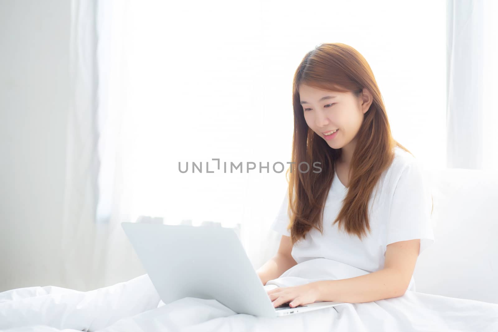 Portrait of beautiful asian young woman setting on bed using laptop computer at bedroom for leisure and relax, freelance with girl working notebook, communication and lifestyle concept.