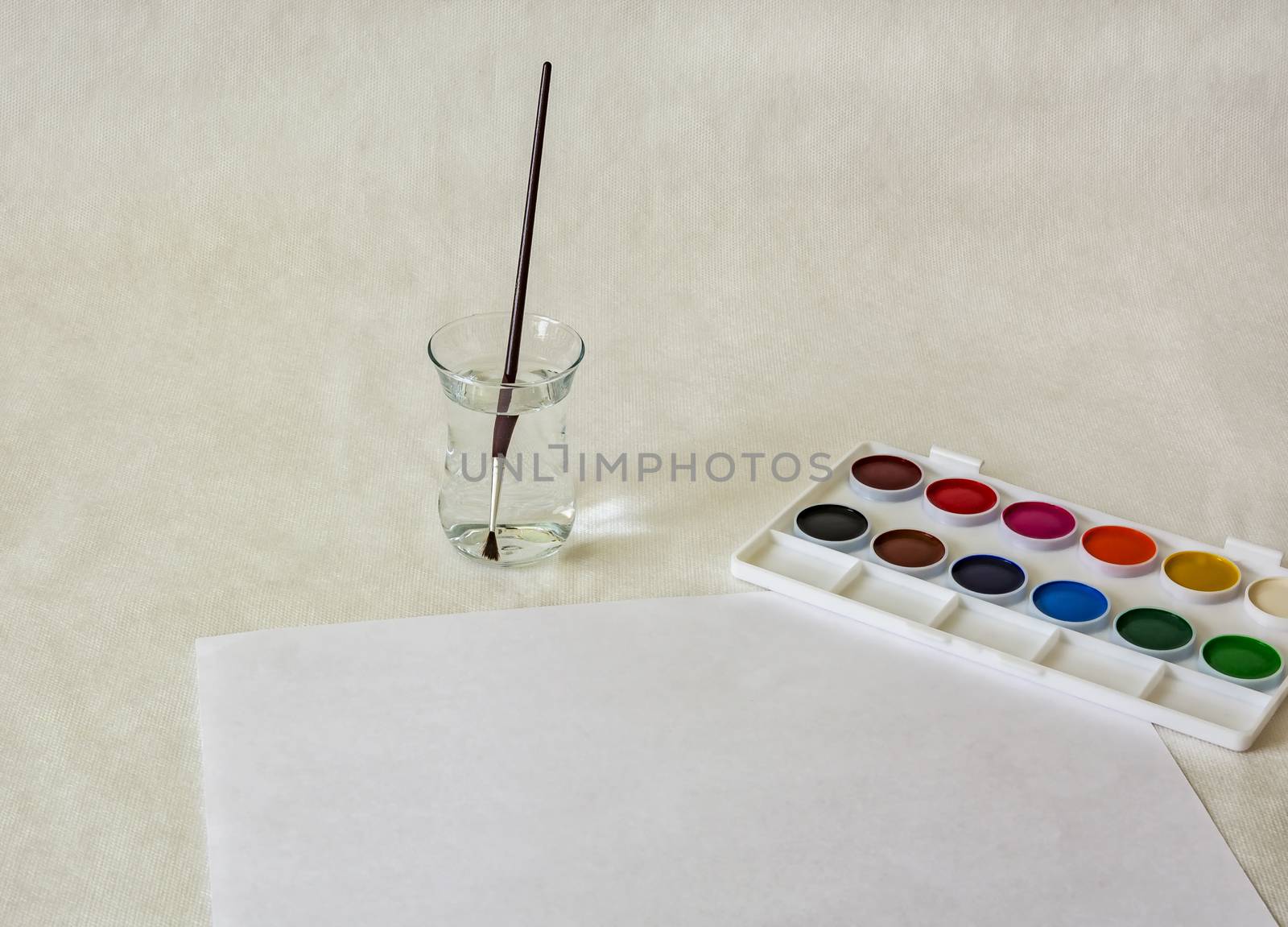 On a light surface there is a set of watercolor paints and there is a glass with water and a brush