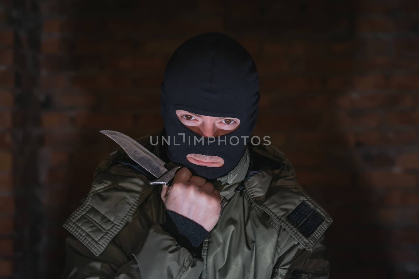 A gangster in a black mask with a knife became bored of standing against a brick wall in the dark