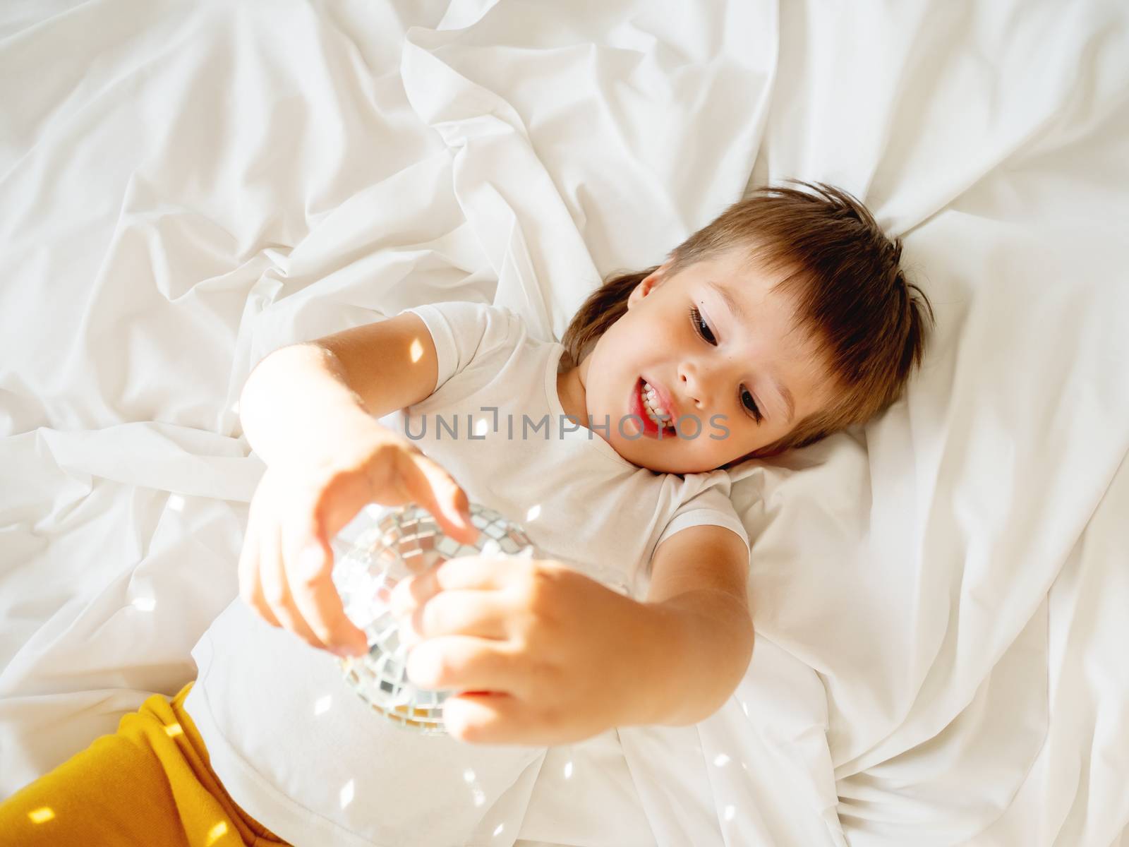 Little boy plays with sparkling mirror disco ball in bed and lau by aksenovko