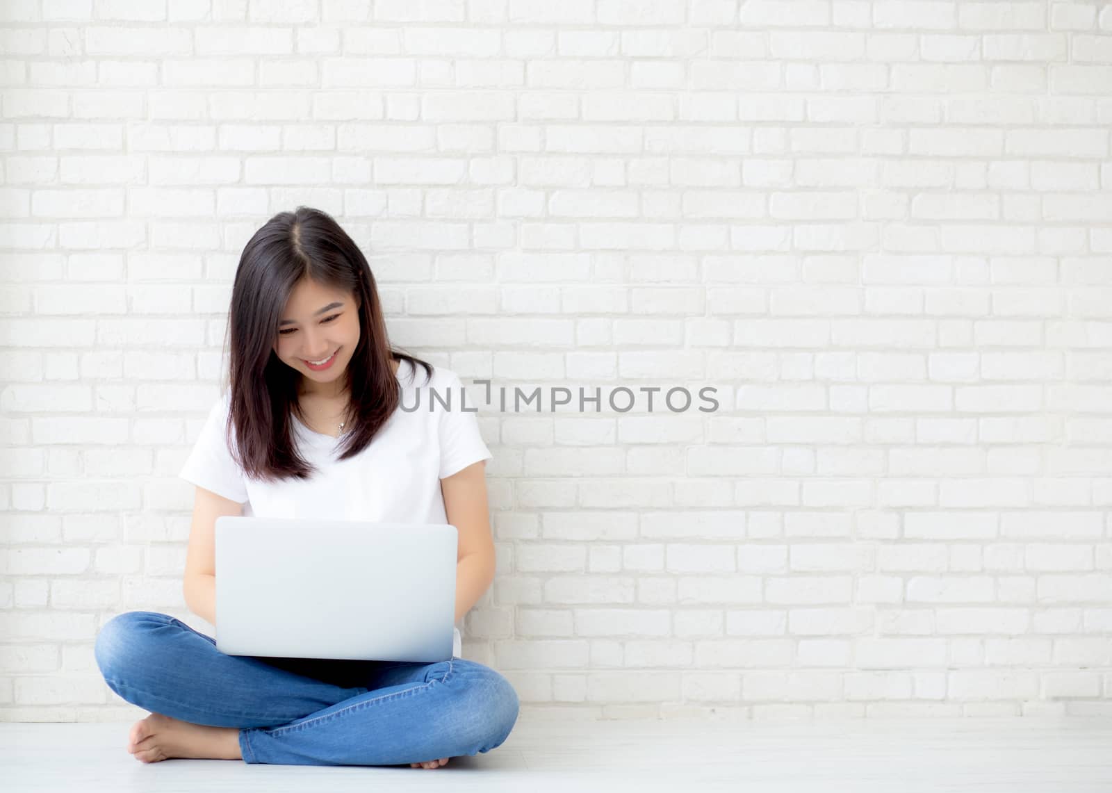 Beautiful of portrait asian young woman working online laptop sitting on floor brick cement background, freelance girl using notebook computer with connect to internet for distance job, business and lifestyle concept.
