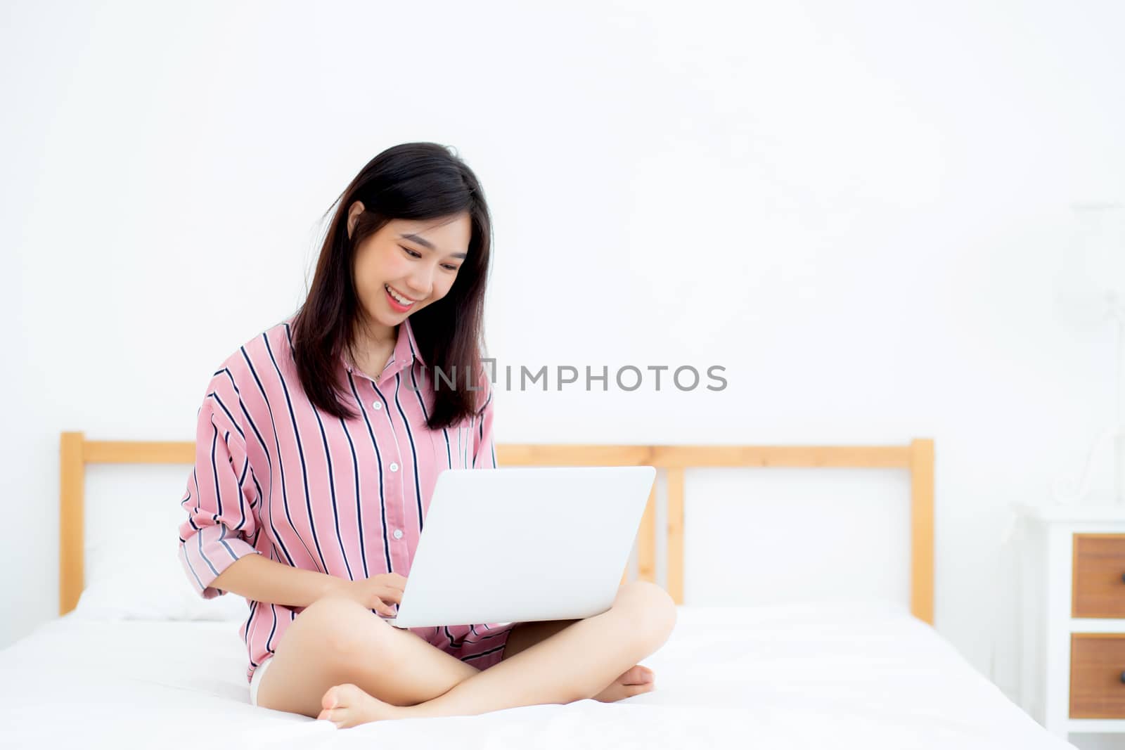 Portrait of beautiful asian young woman setting on bed using laptop computer at bedroom for leisure and relax, freelance with girl working notebook, communication and lifestyle concept.