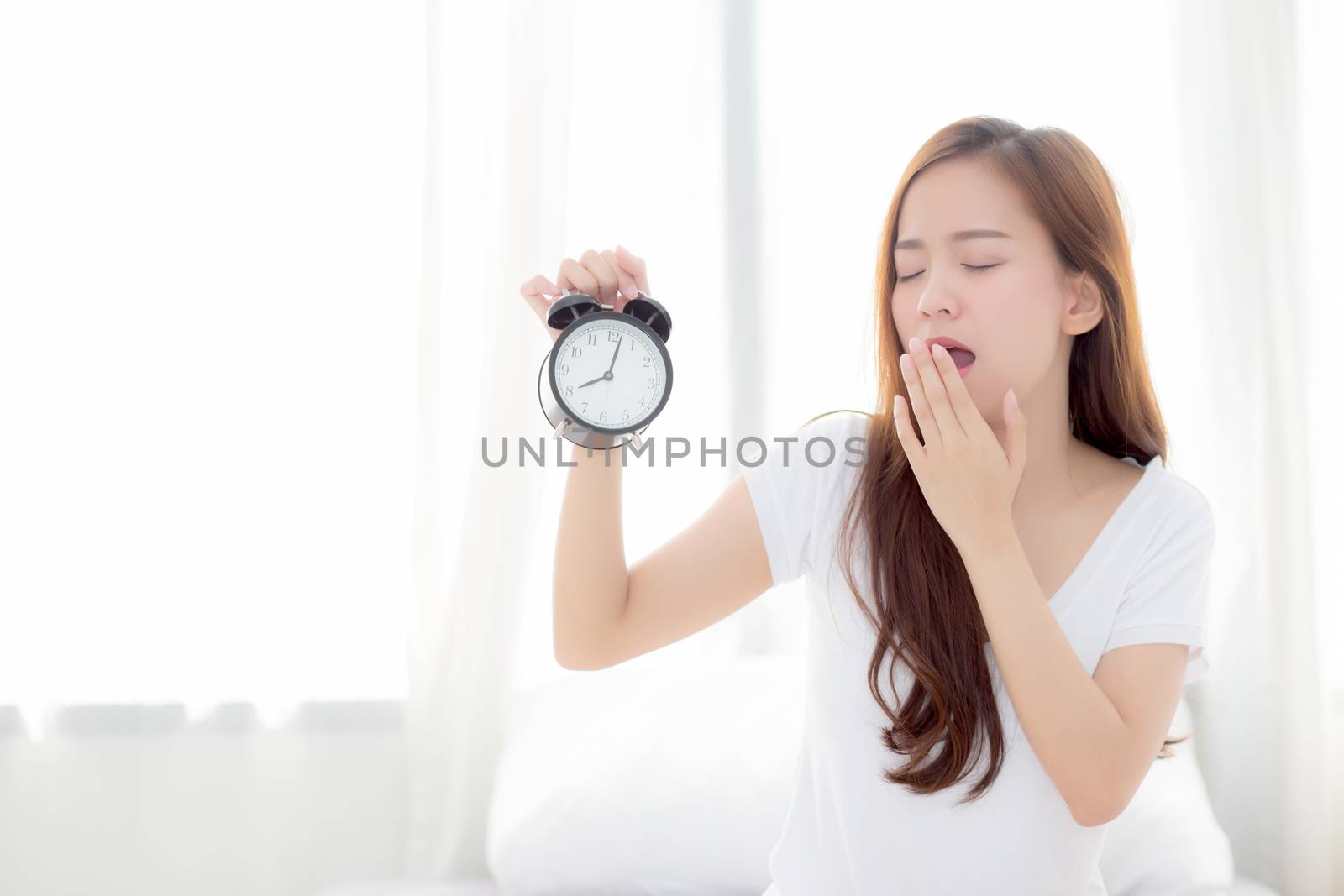 Beautiful young asian woman wake up in morning yawn and annoyed alarm clock holding hand, girl standing hurry wake late with appointment with curtain background on bedroom, lifestyle concept.