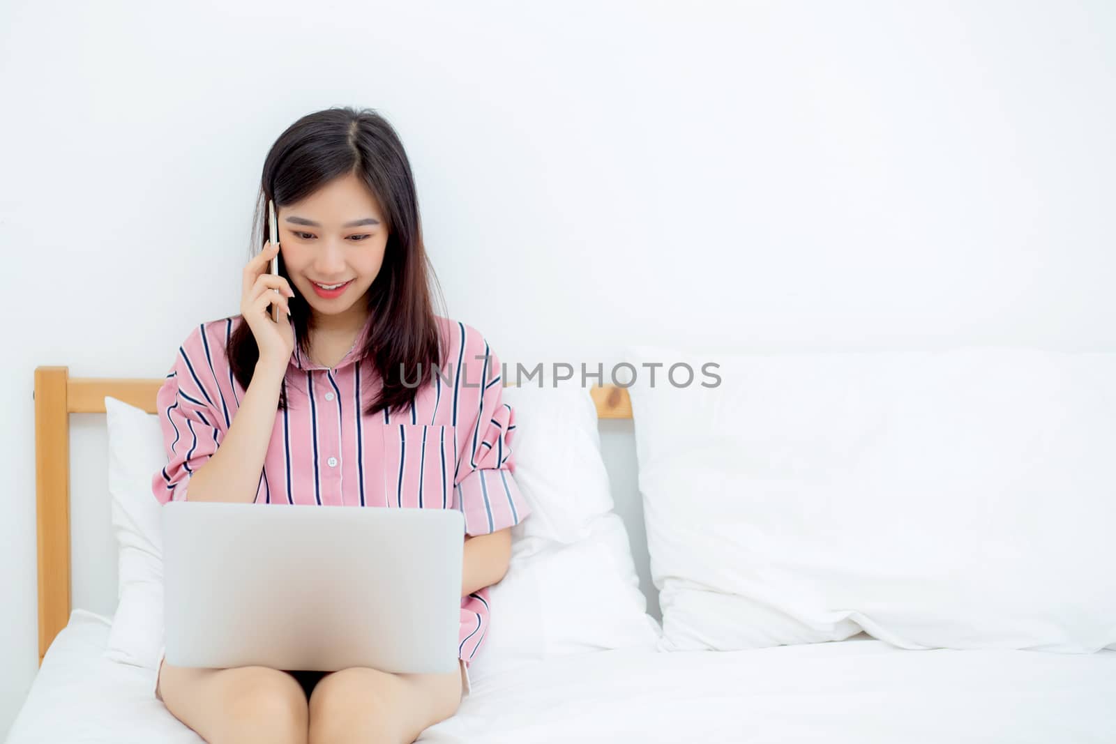 Portrait of beautiful asian young woman setting on bed using laptop computer and talk smart phone at bedroom for leisure and relax, freelance with girl working notebook and calling telephone, communication and lifestyle concept.