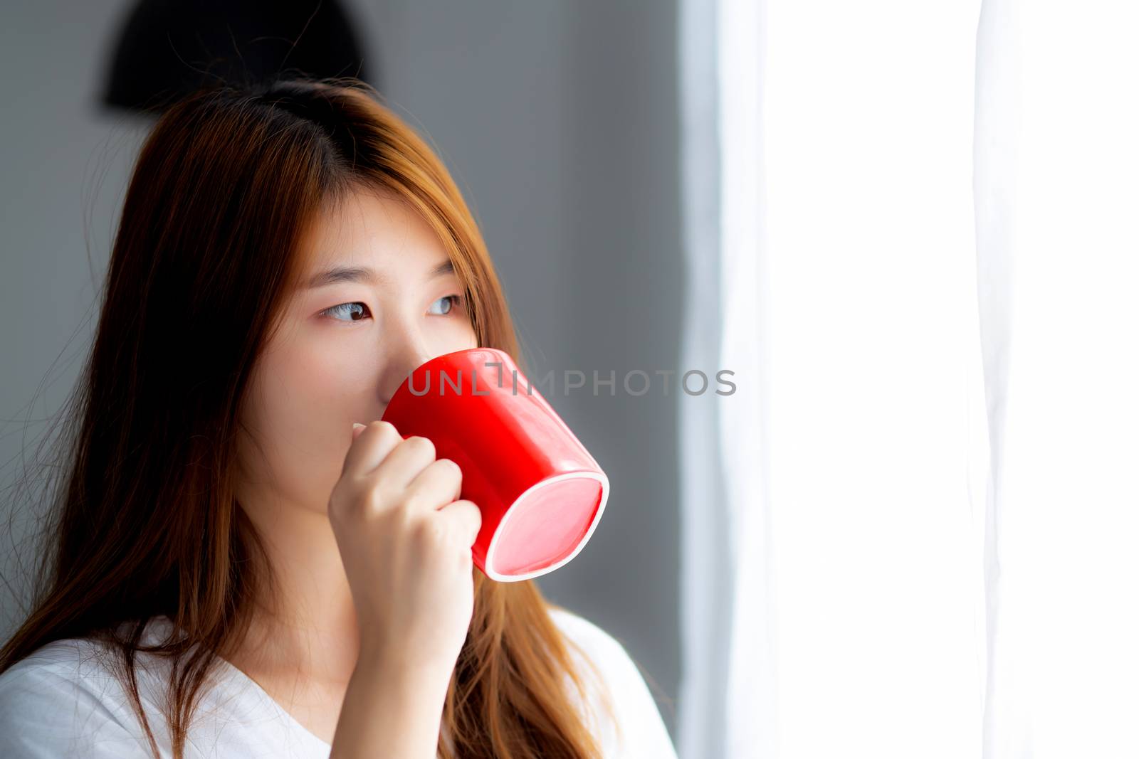 Beautiful of portrait young asian woman with drink acup of coffee standing curtain window background in bedroom, girl relax in morning at home, lifestyle concept.
