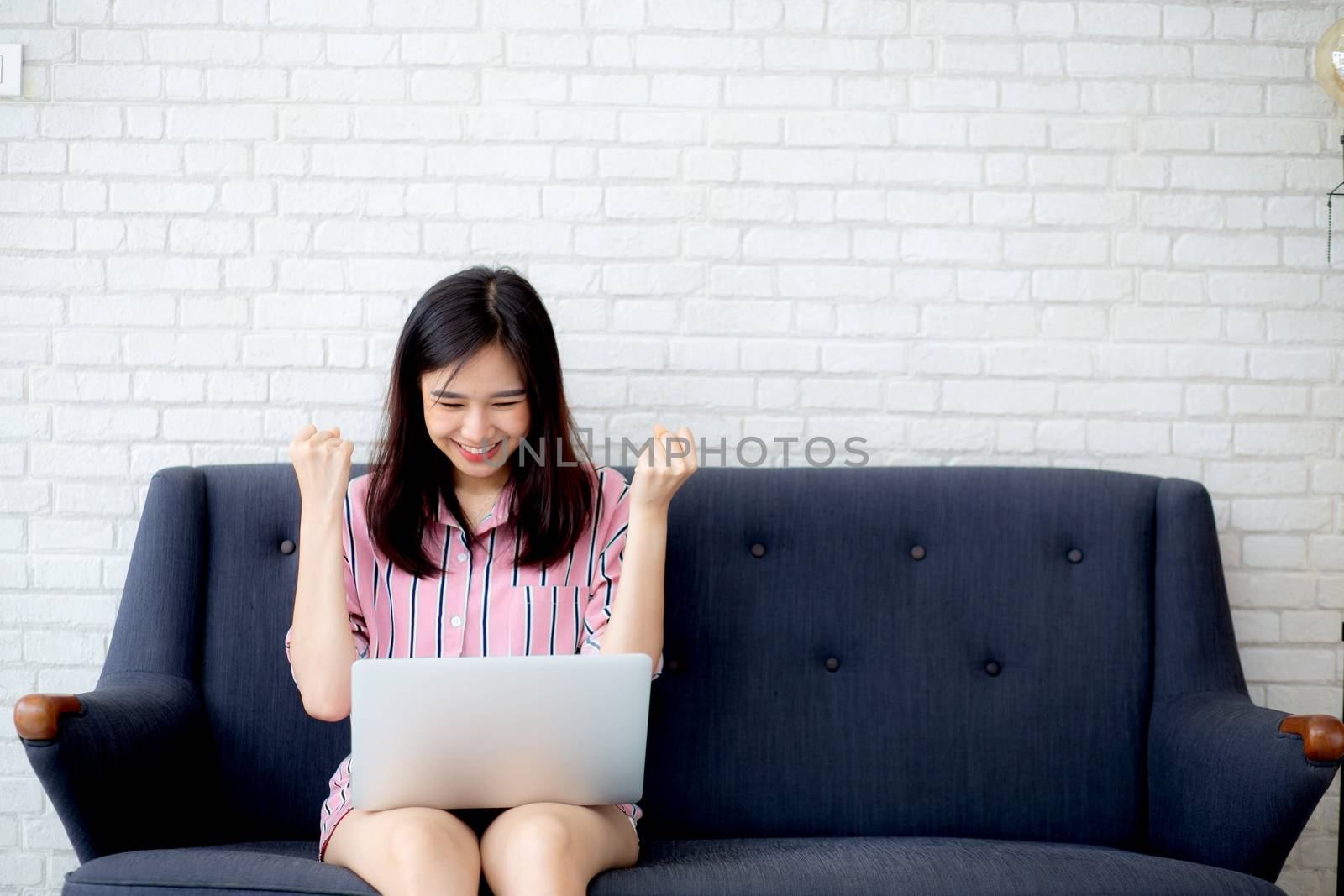 Beautiful of portrait asian young woman excited and glad of success with laptop on sofa cement background, girl working living room at home, career freelance business concept.