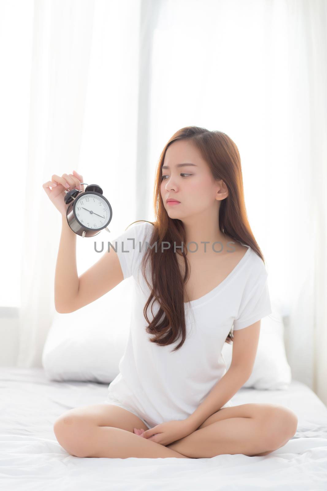 Beautiful young asian woman wake up in morning annoyed alarm clock holding hand, girl sitting hurry wake late with appointment with curtain background on bedroom, lifestyle concept.