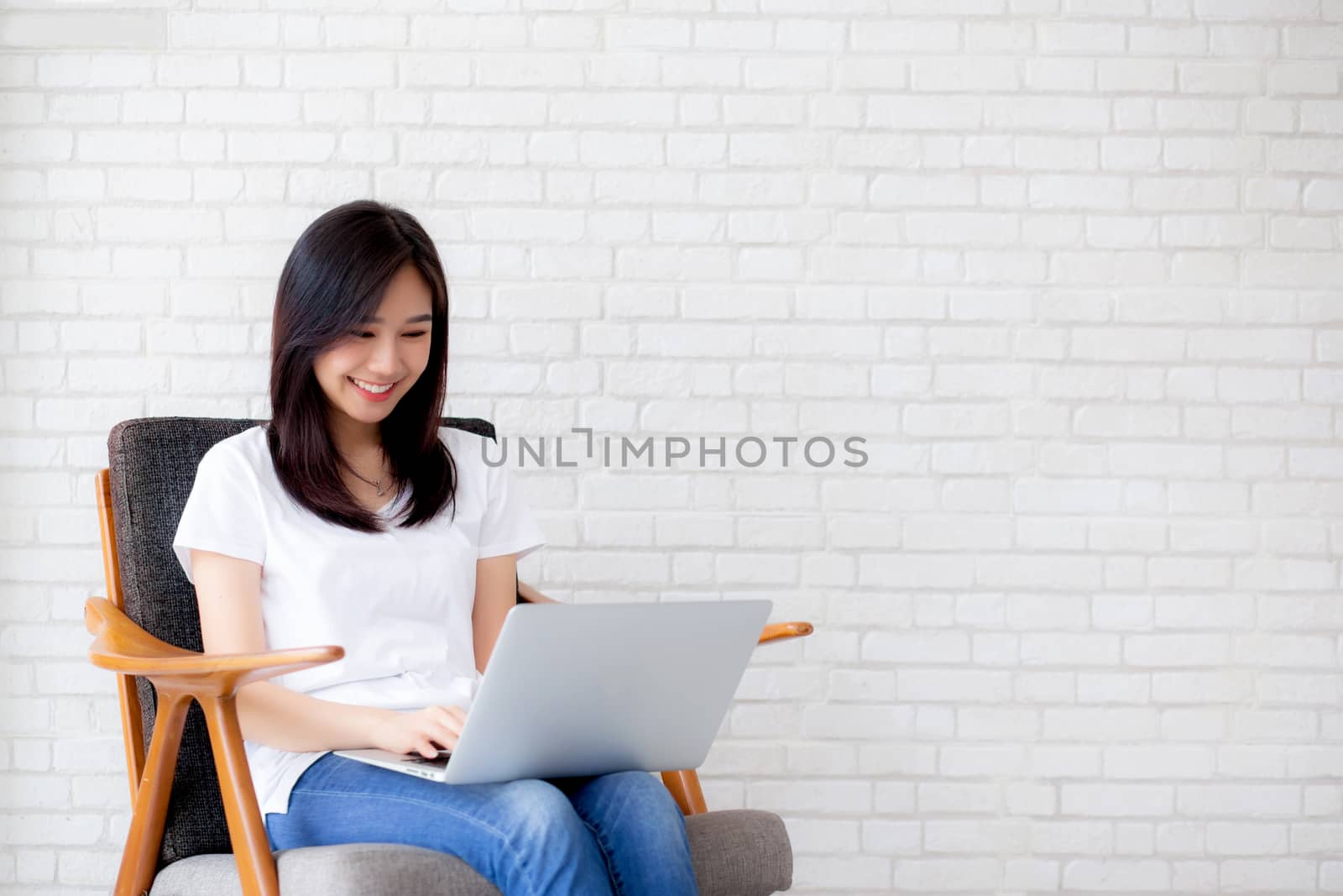 Beautiful of portrait asian young woman working online laptop sitting on chair on cement brick white background, girl using notebook computer, business concept.