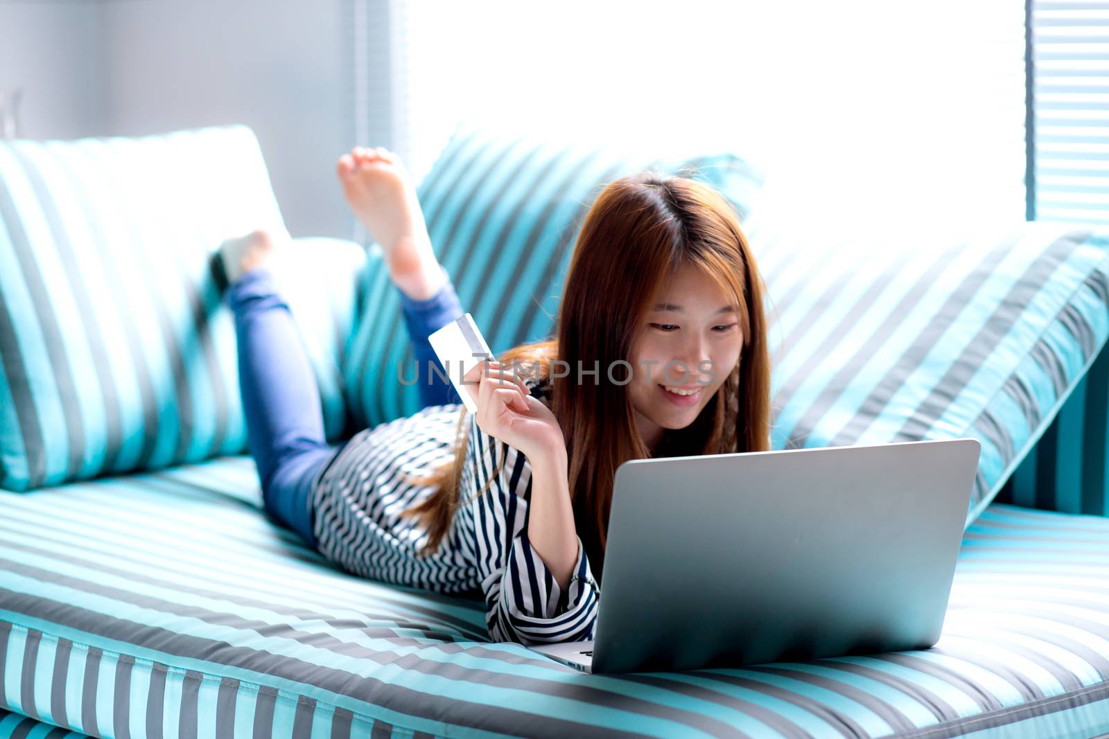 Beautiful of portrait young asian woman lying users credit card with laptop, Content girl shopping online and payment with notebook computer on sofa, lifestyle concept.