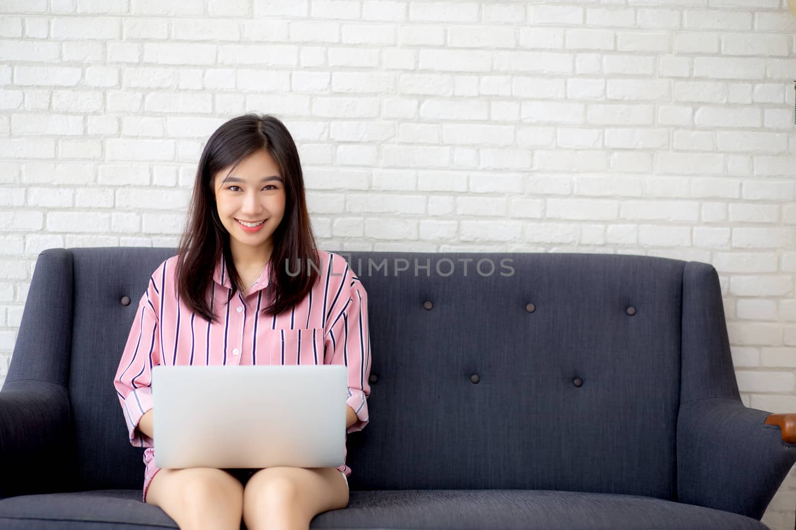 Beautiful of portrait asian young woman working online laptop sitting on sofa at living room, girl using notebook computer with connect to internet for distance job, business and success concept.