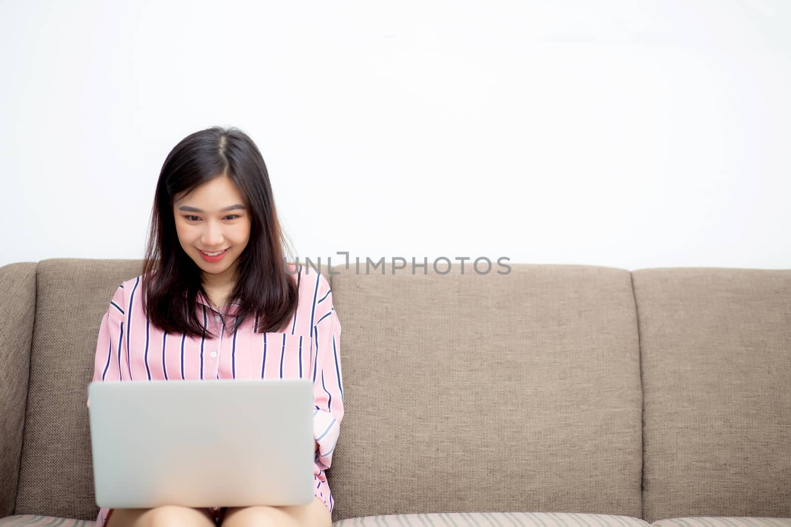 Beautiful of portrait asian young woman working online laptop sitting on sofa at living room, girl using notebook computer with connect to internet for distance job, business and success concept.