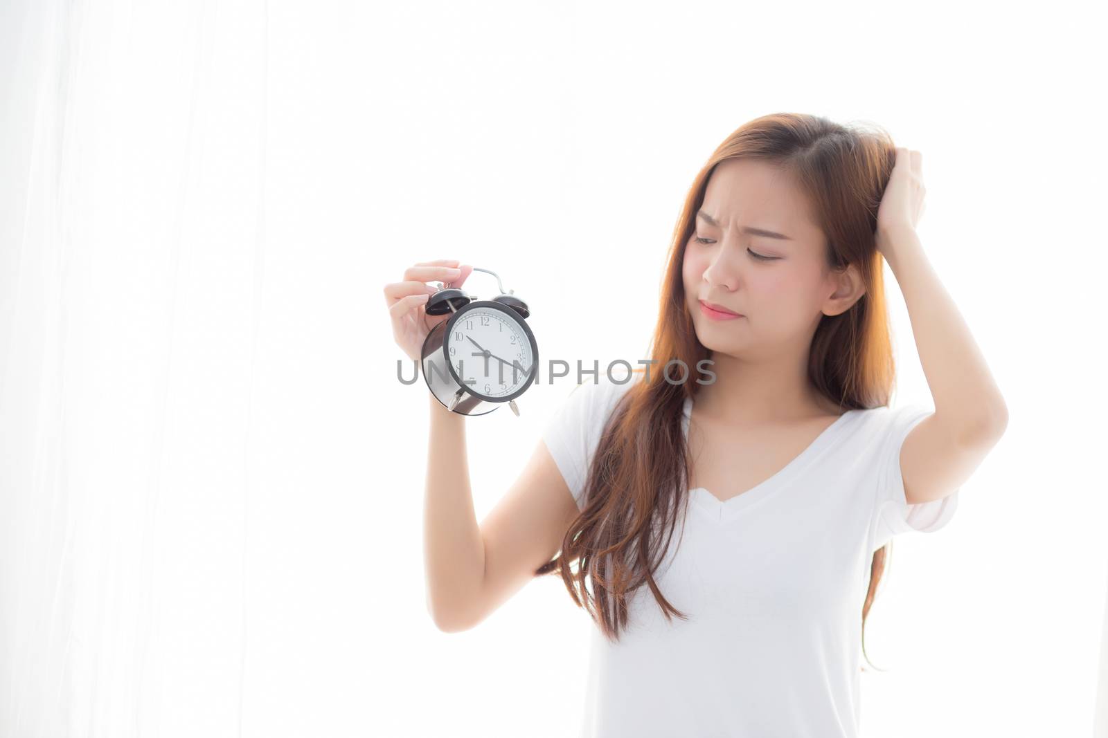 Beautiful young asian woman wake up in morning annoyed alarm clock holding hand, girl standing hurry wake late with appointment with curtain background on bedroom, lifestyle concept.