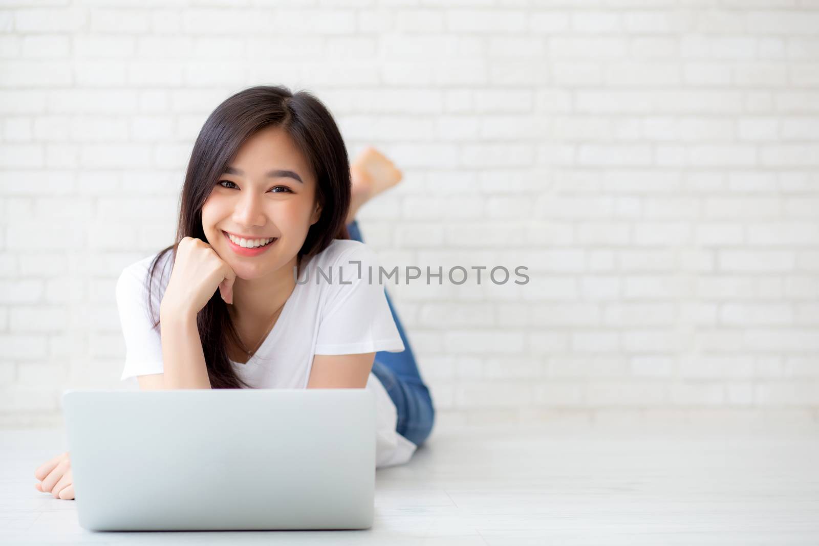 Beautiful of portrait asian young woman working online laptop lying on floor brick cement background, freelance girl using notebook computer with connect to internet for distance job, business and lifestyle concept.