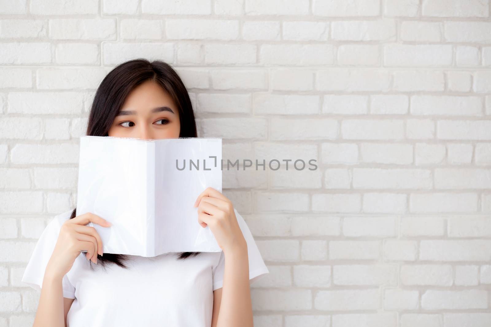 beautiful portrait young asian woman happy hiding behind open the book with cement or concrete background, girl standing reading for learning, education and knowledge concept.