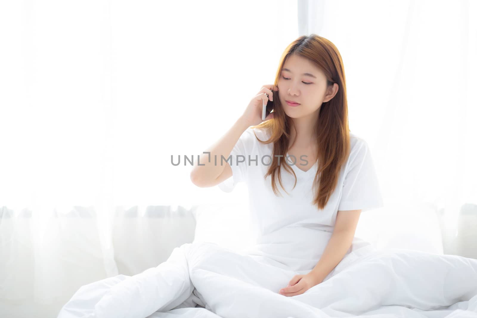 Beautiful young asian woman smiling sitting and relax on the bed in the morning, girl using mobile smart phone talking enjoy at bedroom, communication concept.