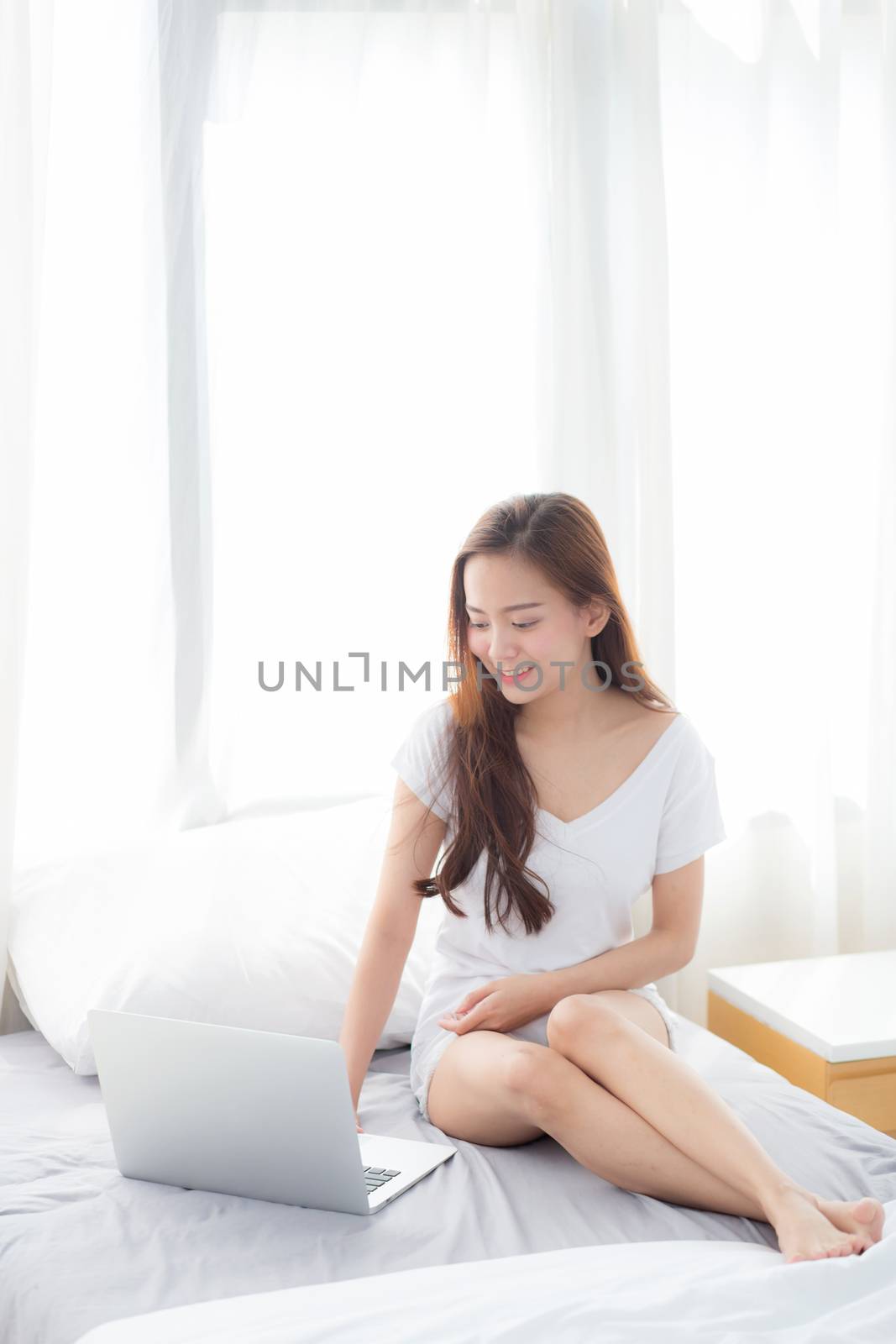 Portrait of beautiful asian young woman setting on bed using laptop computer at bedroom for leisure and relax, freelance with girl working notebook, communication and lifestyle concept.