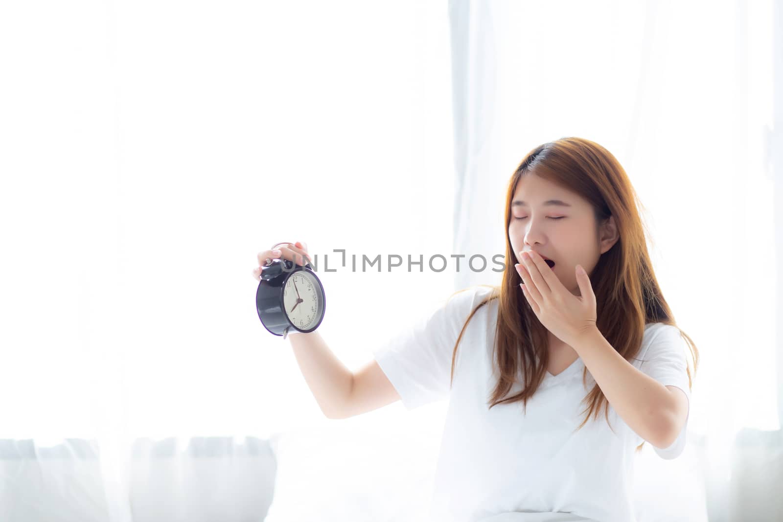 Beautiful young asian woman yawn wake up in morning annoyed alarm clock holding hand, standing hurry wake late with appointment with curtain background on bedroom, lifestyle concept.