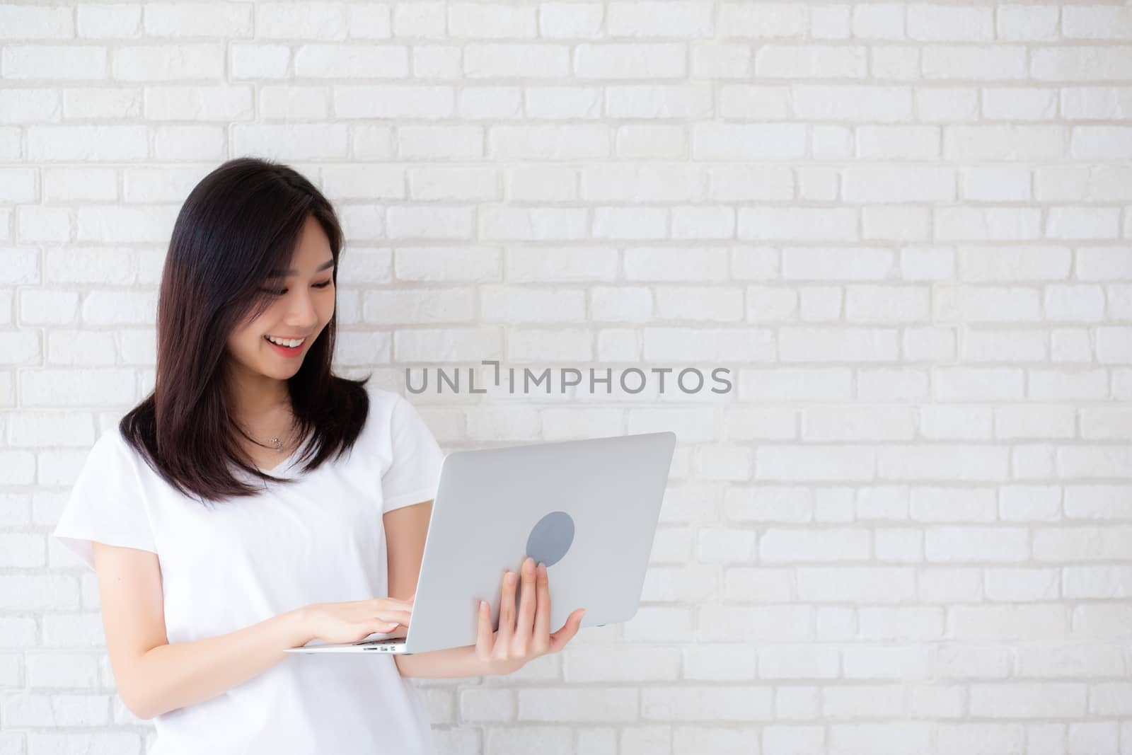 Beautiful portrait asian young woman smile using laptop standing at workplace on cement concrete background, girl happy with computer internet online, lifestyle and freelance business concept.