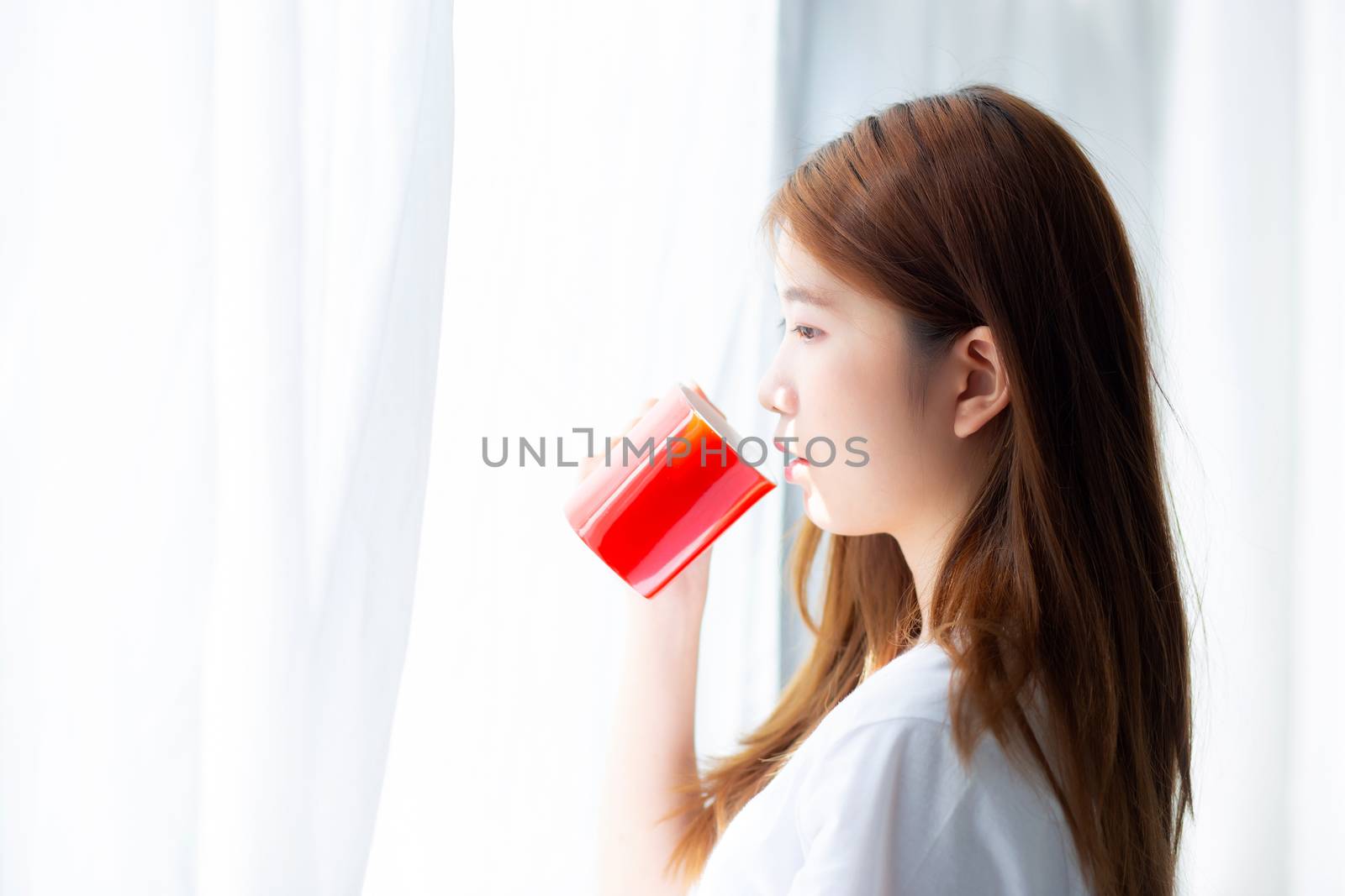 Beautiful of portrait young asian woman with drink acup of coffee standing curtain window background in bedroom, girl relax in morning at home, lifestyle concept.