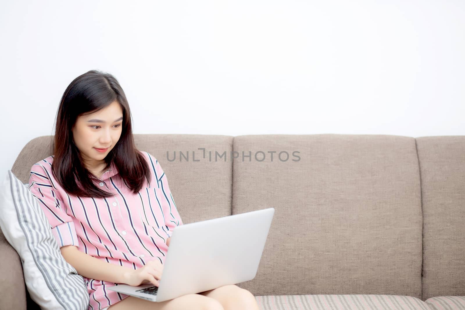 Beautiful of portrait asian young woman working online laptop with smile and happy sitting on sofa at living room, girl using notebook computer with connect to internet, business and success concept.