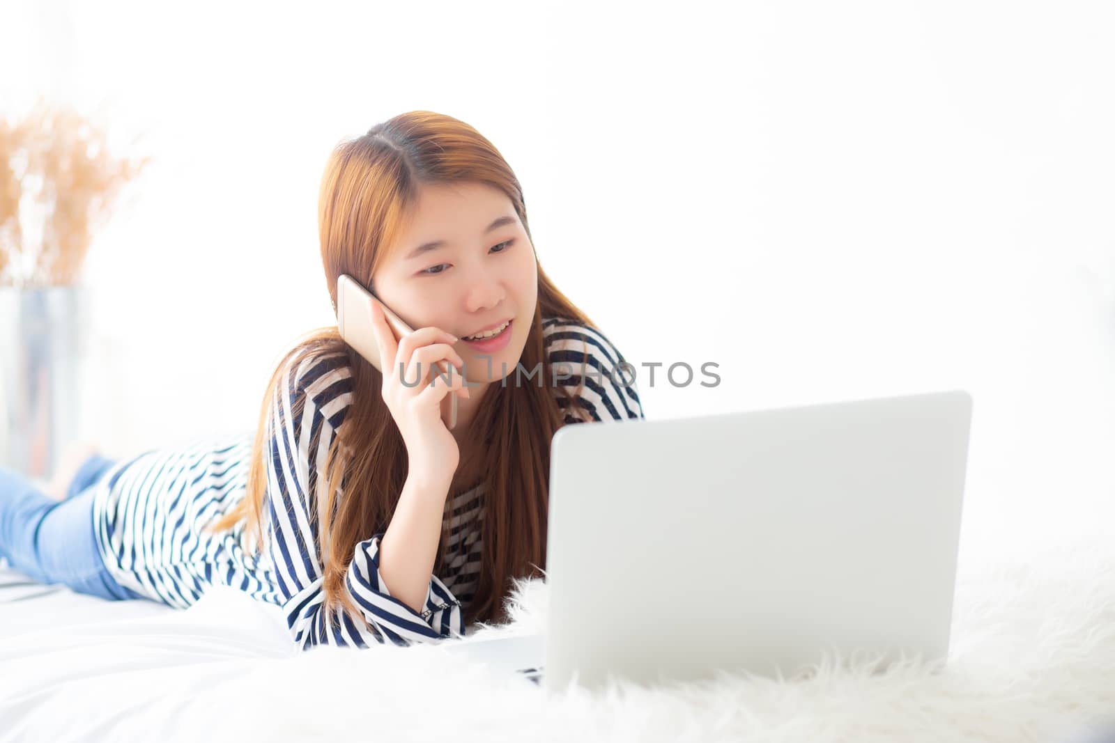 Beautiful asian young woman lying on bed using laptop and talking mobile smart phone at bedroom for leisure and relax, freelance with girl working notebook, communication concept.