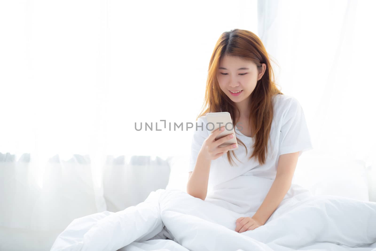 Beautiful young asian woman smiling lying relax on the bed in the morning, girl using touch and talking mobile smart phone enjoy, communication and social network concept.