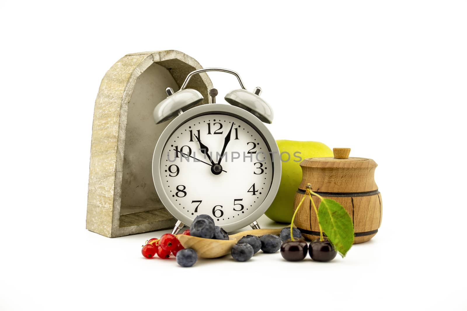 Culinary still life with old-fashioned alarm clock, apple, jar of honey, cherries, red currants and blueberries over white background with copy space