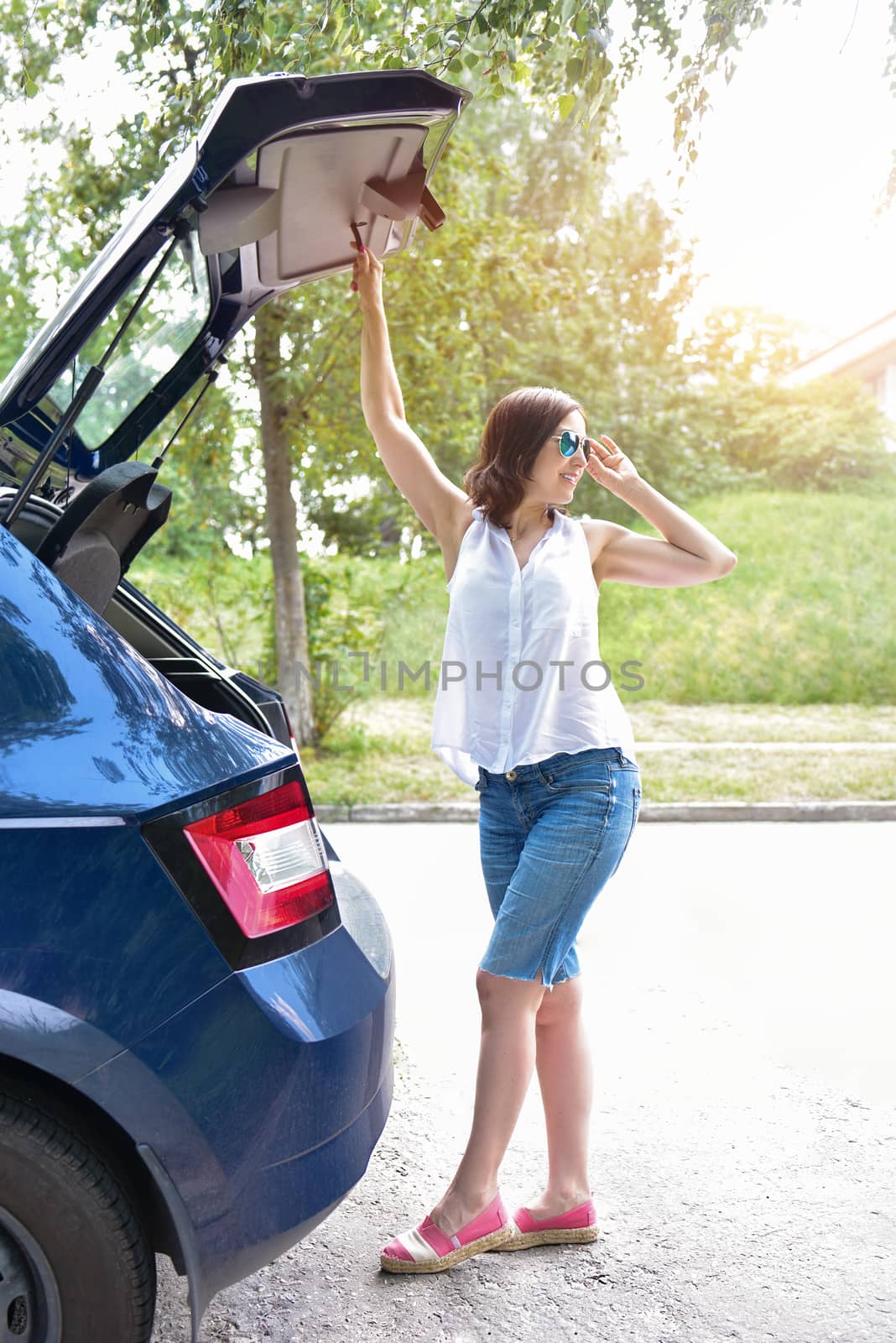 Smiling Caucasian woman in a white blouse and denim shorts putting her stuff bags into the car trunk