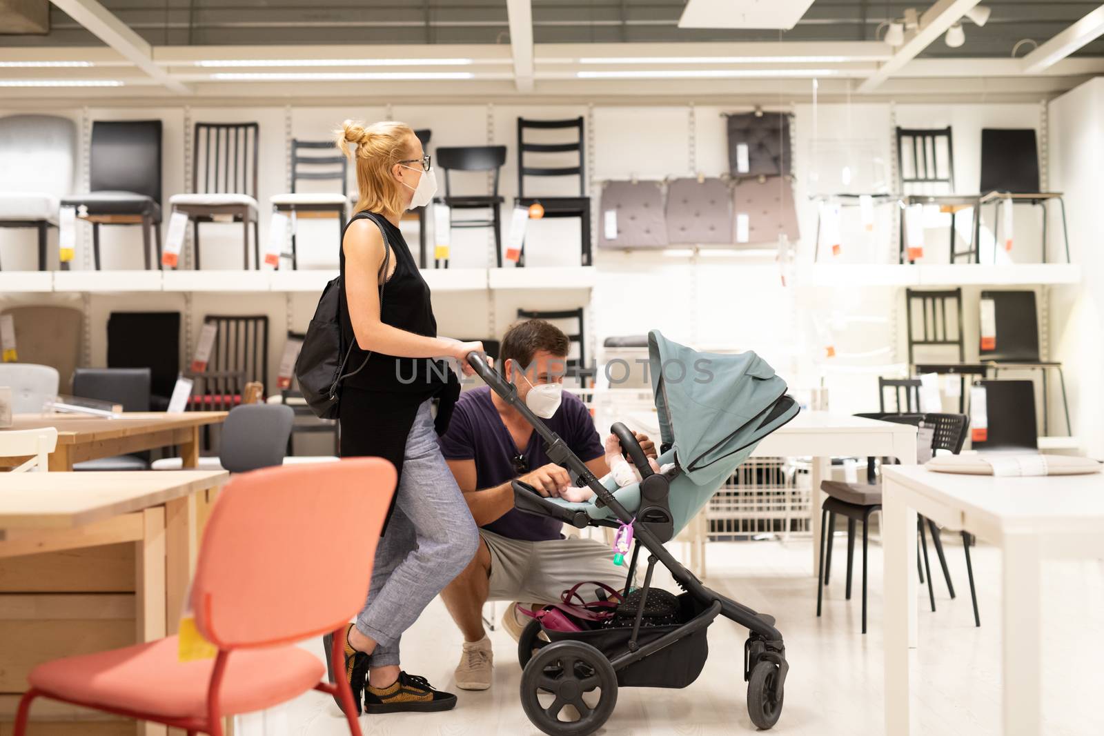 Young couple with newborn in stroller shopping at retail furniture and home accessories store wearing protective medical face mask to prevent spreading of corona virus when shops reopen. by kasto