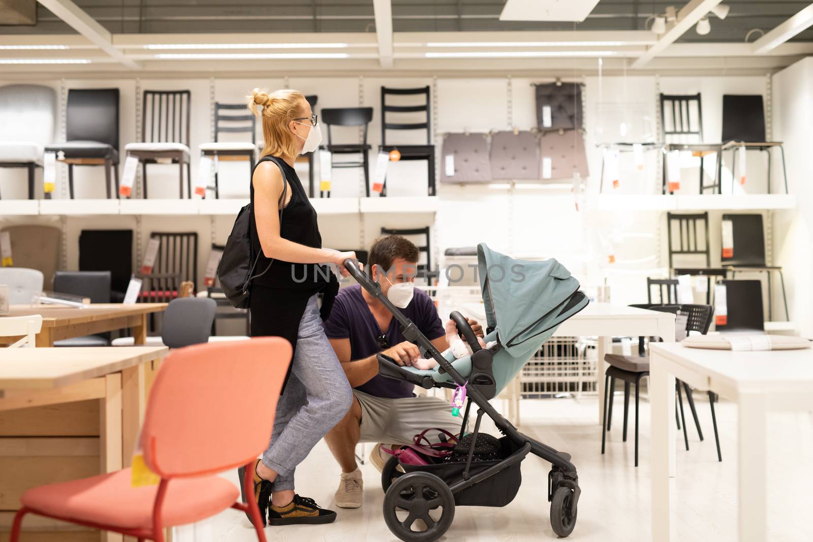 Young family with newborn in stroller shopping at retail furniture and home accessories store wearing protective medical face mask to prevent spreading of corona virus when shops reopen.