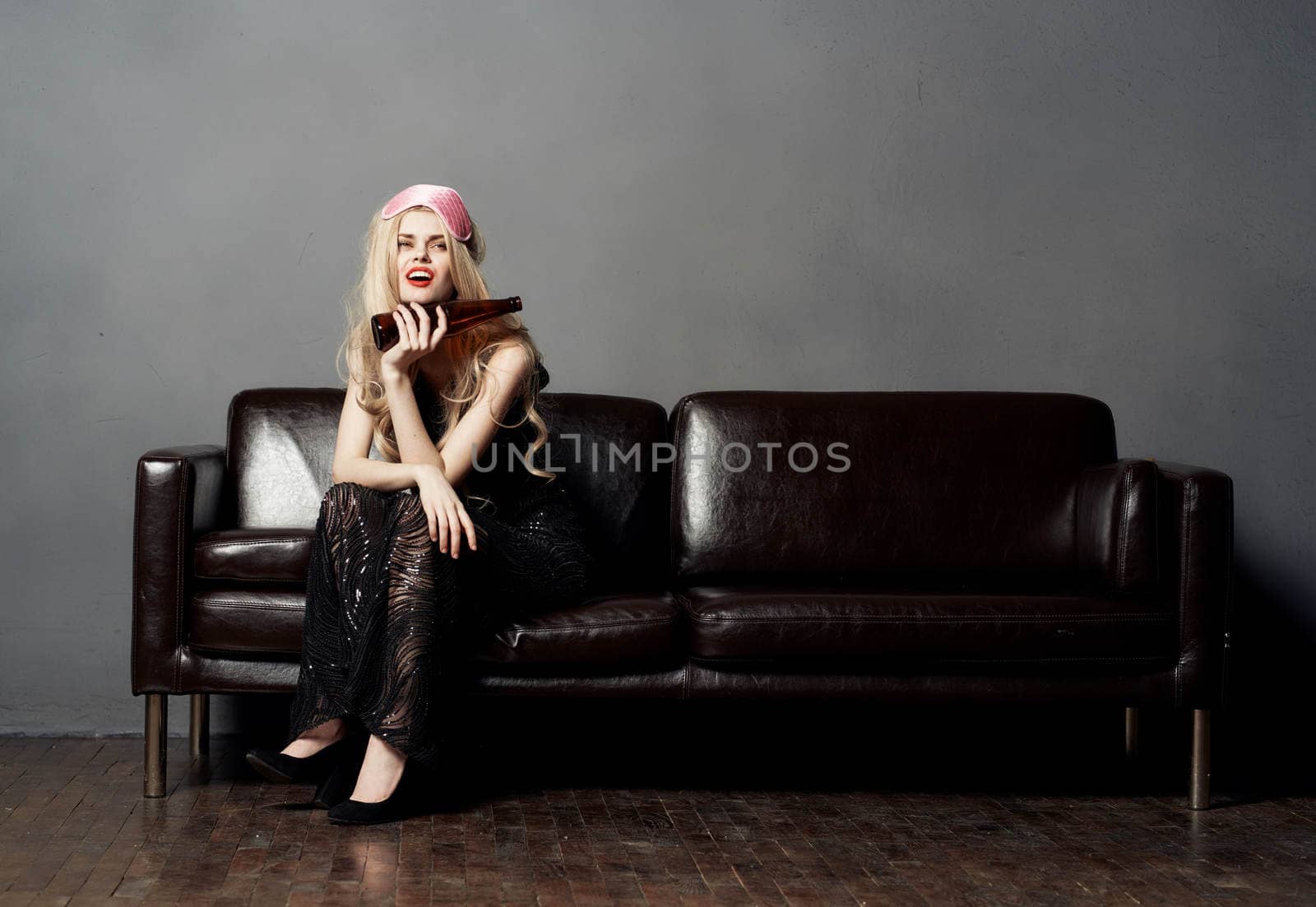 A blonde woman with a bottle of beer on a leather sofa in a black dress. High quality photo