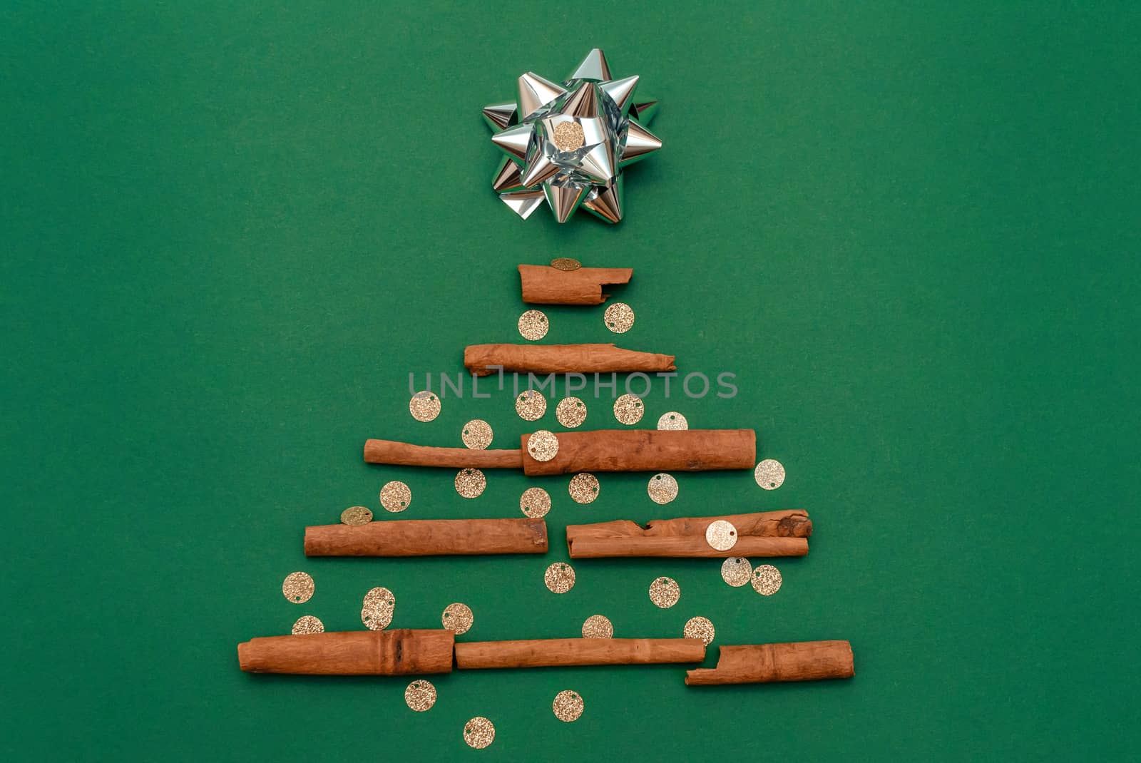 Christmas tree made of cinnamon sticks with a silver gift bow on a green background with golden confetti, copy space. Flat lay. New year card and template for design.
