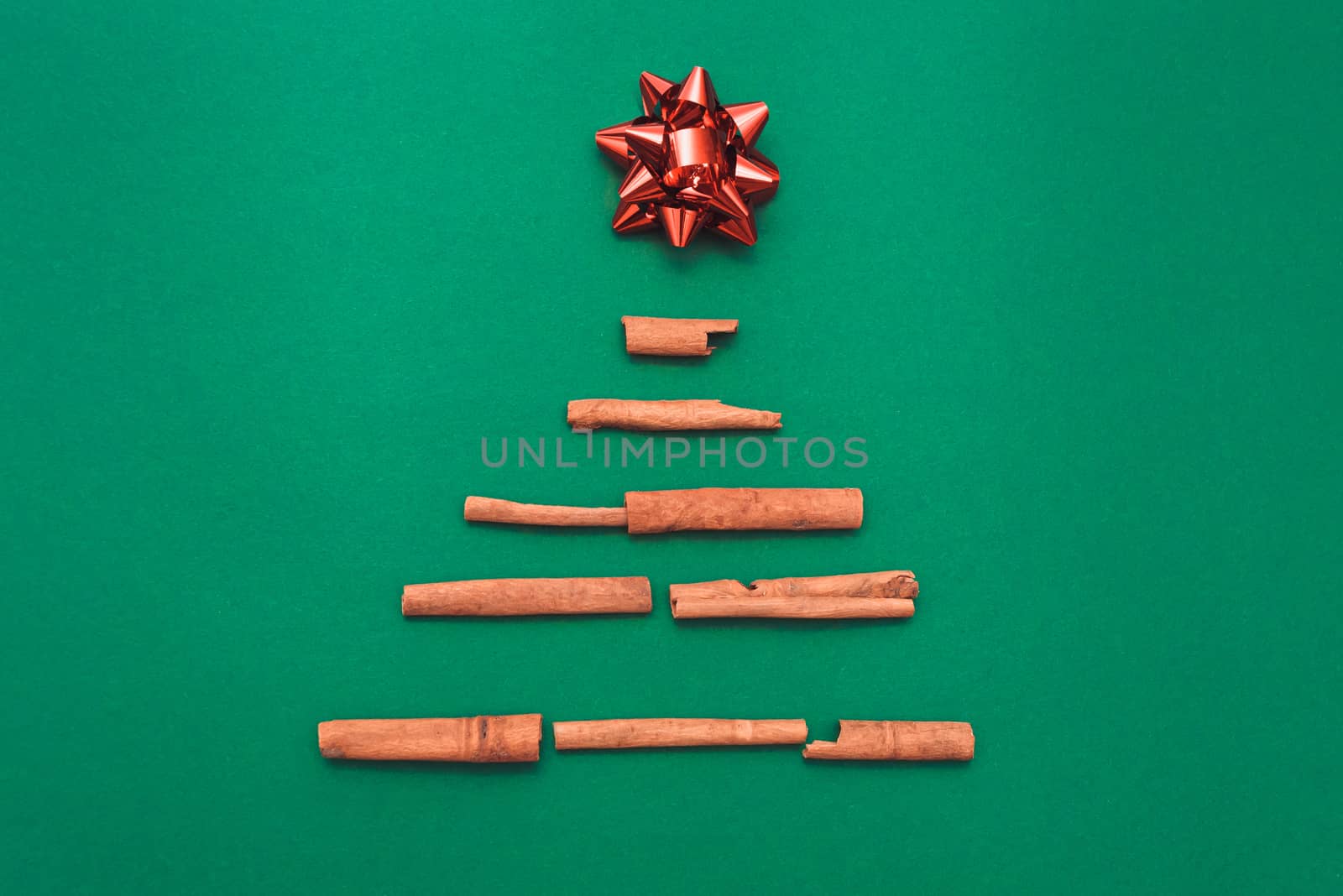 Christmas tree made of cinnamon sticks with a red gift bow on a green background, copy space. Flat lay. New year card and template for design.