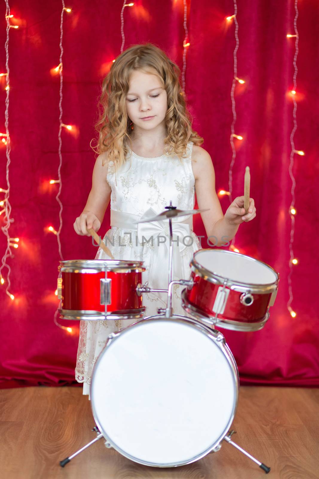 Little smiling girl plays on drums on red background with new year garlands.