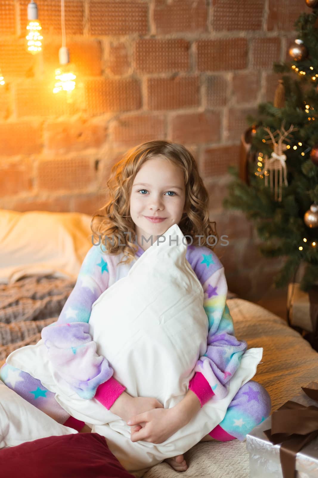 Cute little girl sits on the bed and huggs pillows with background of christmas tree behaind.